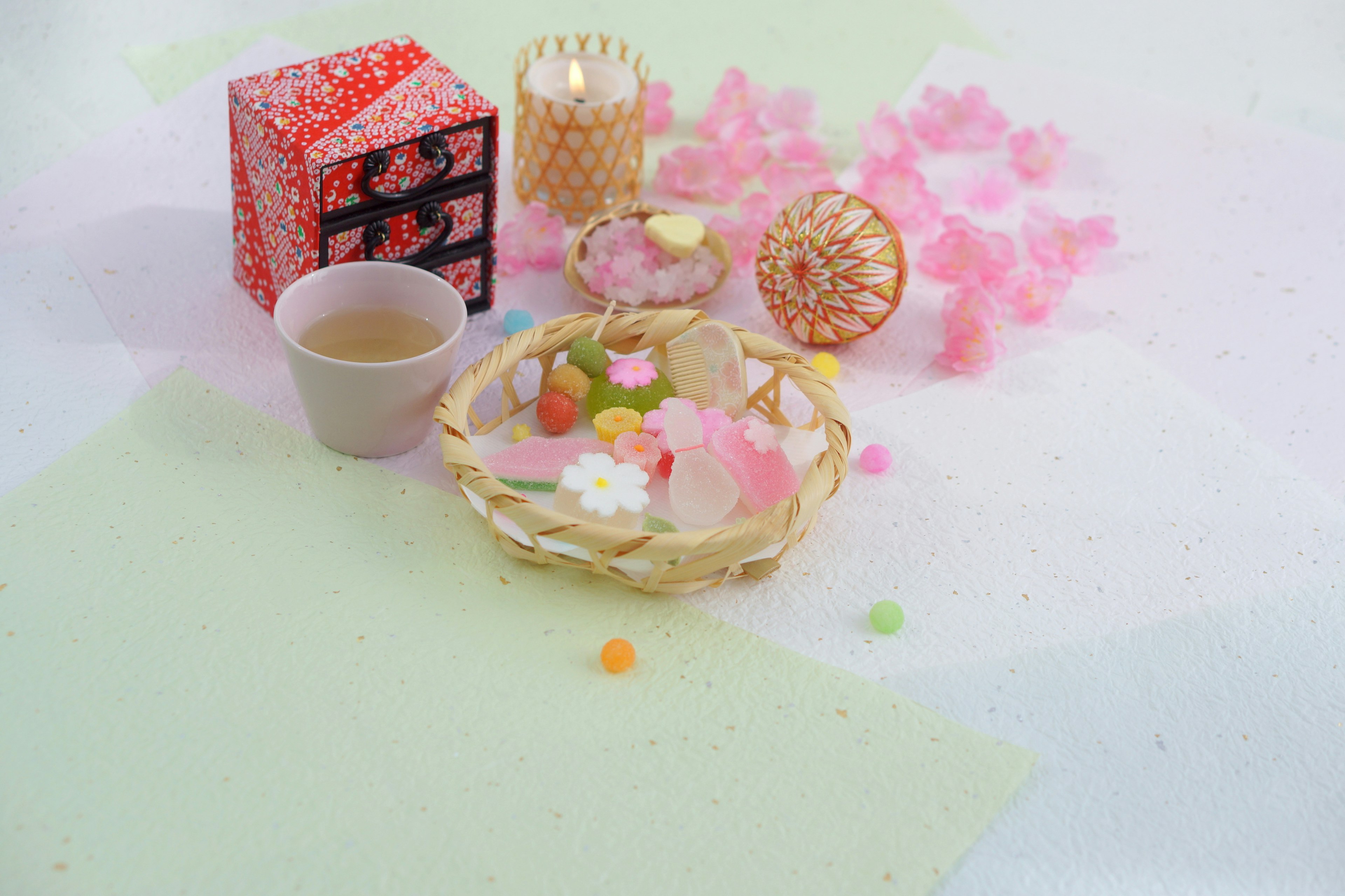 Una pequeña canasta llena de caramelos coloridos rodeada de pétalos de cerezo rosa y objetos decorativos