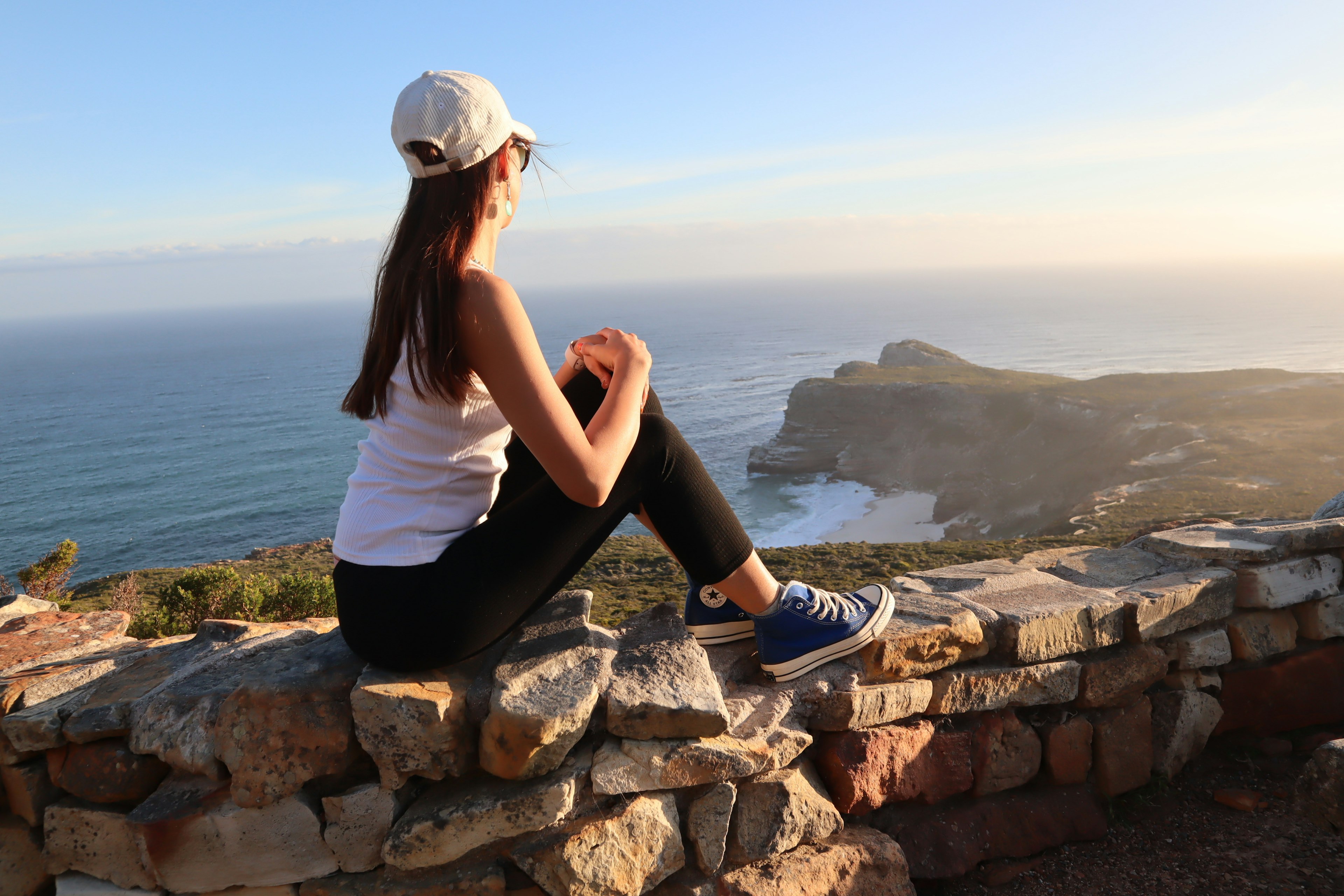 Woman sitting on a stone wall overlooking the ocean wearing a white cap black pants and sneakers