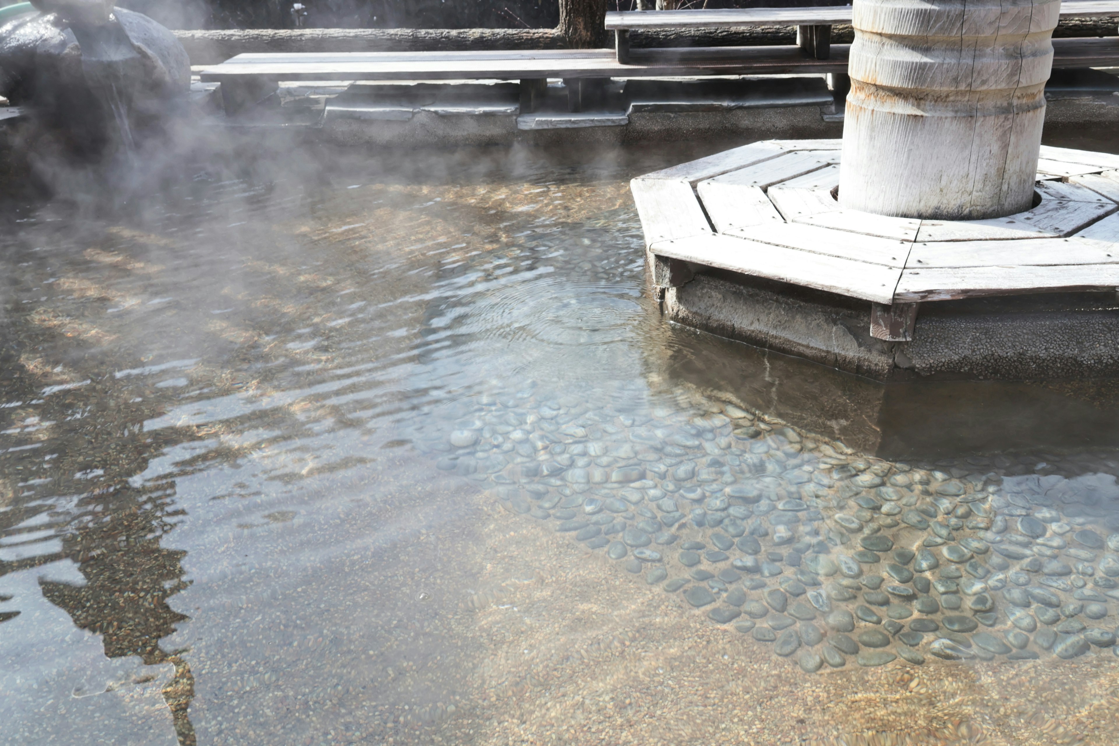 Image of a hot spring with steam rising from the water surface visible stones in shallow water