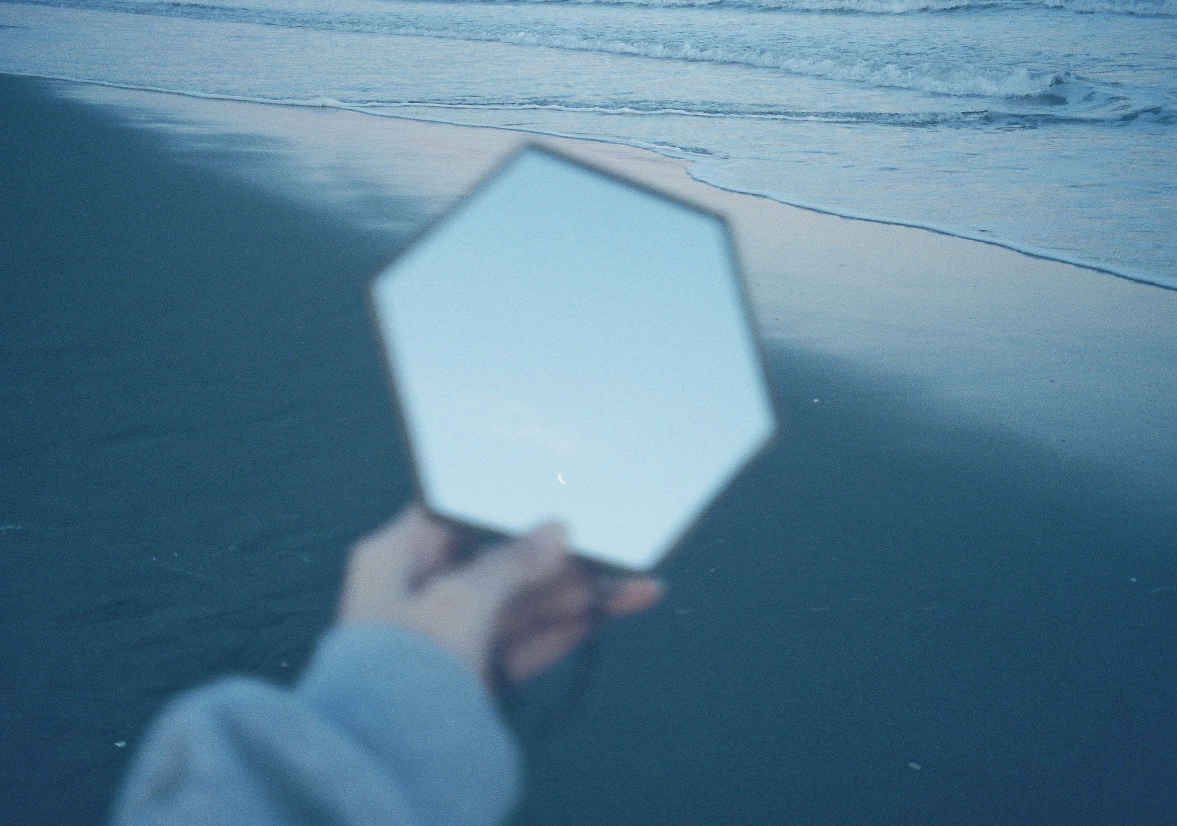 Imagen borrosa de una mano sosteniendo un espejo hexagonal en la playa