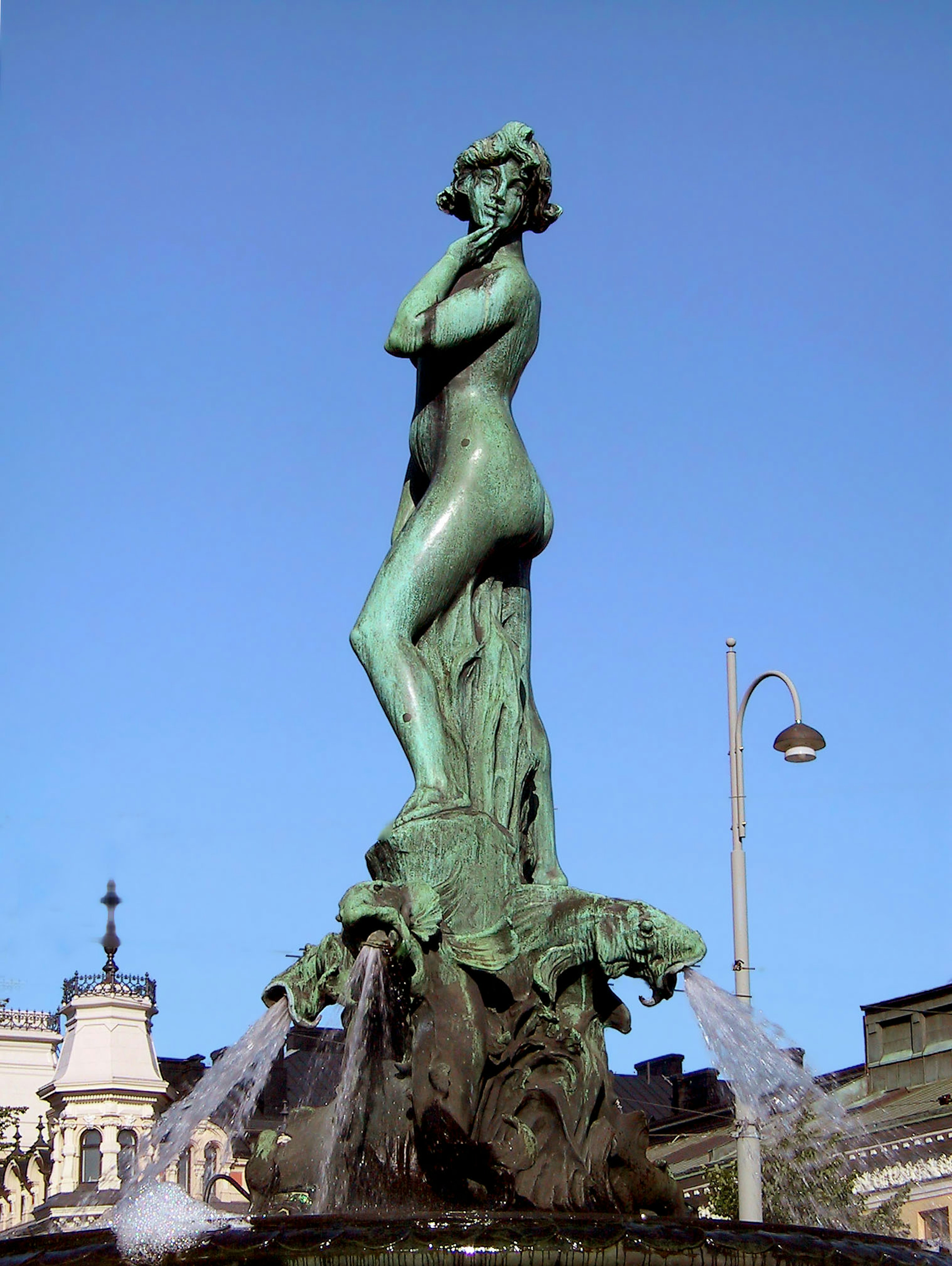 Statue of a nude figure in green bronze with flowing water at the base against a blue sky