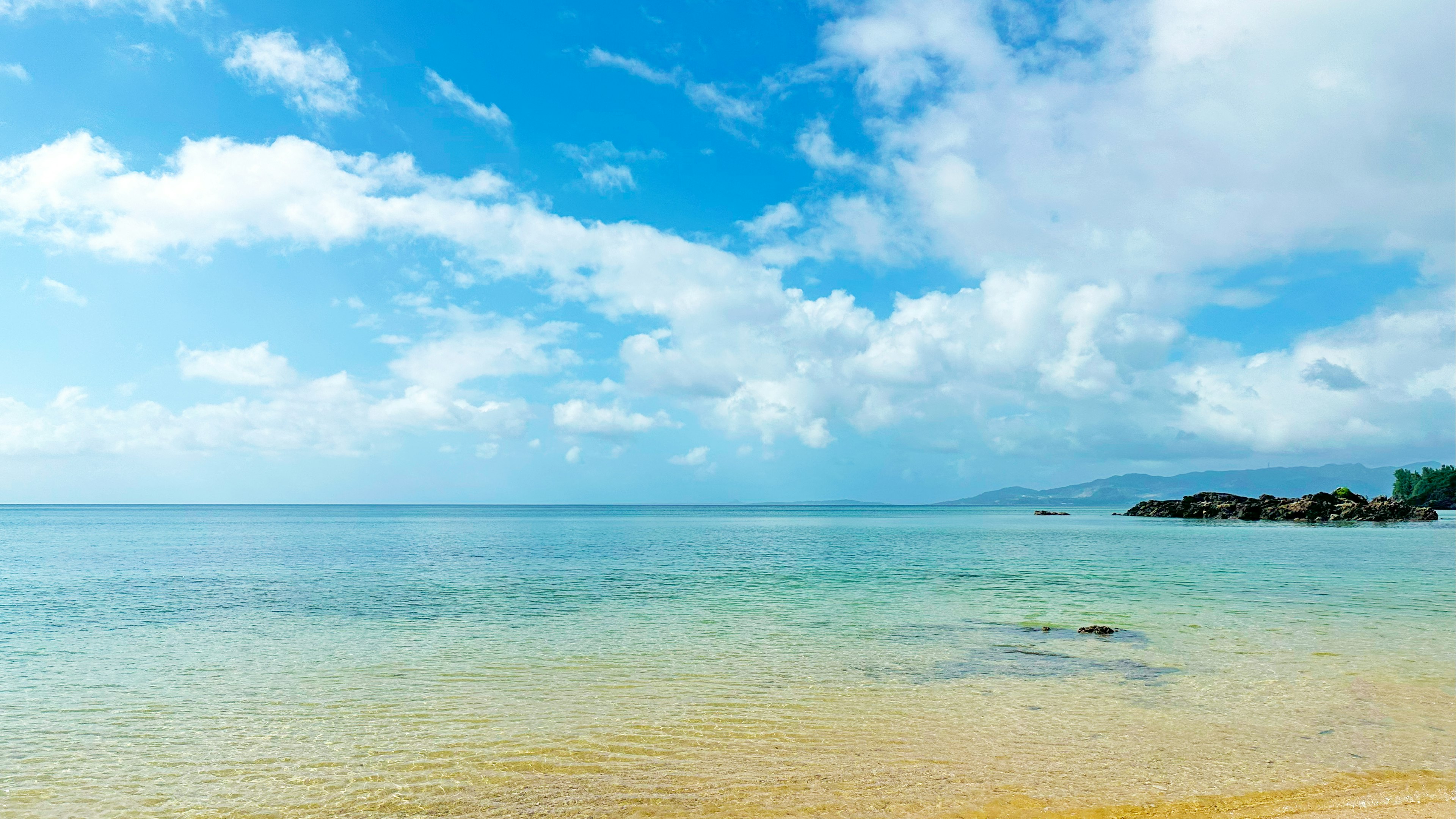 Pemandangan pantai dengan langit biru dan awan putih, ombak tenang dan pantai berpasir