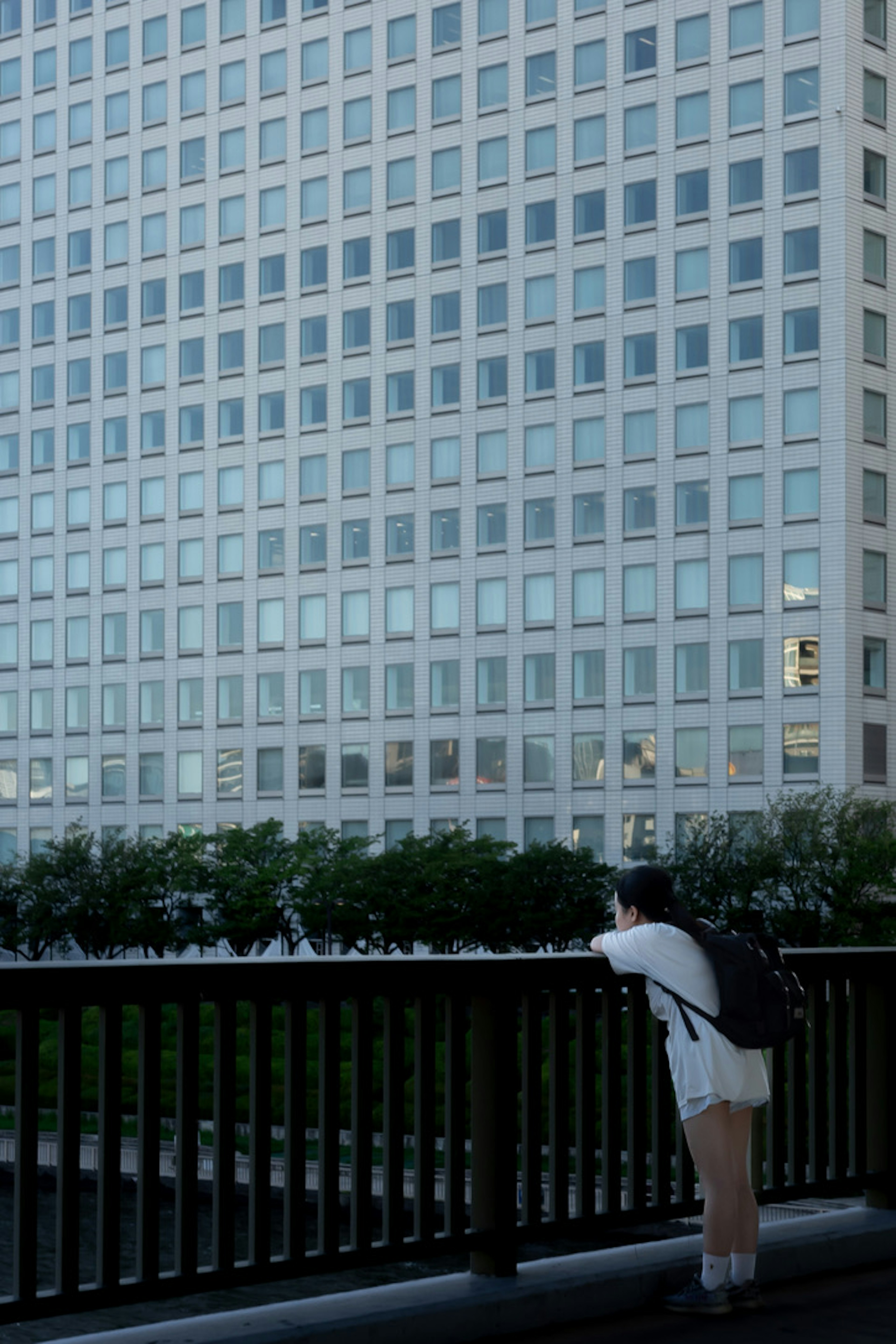 Young woman leaning on railing in front of tall building blue sky and greenery reflected in windows