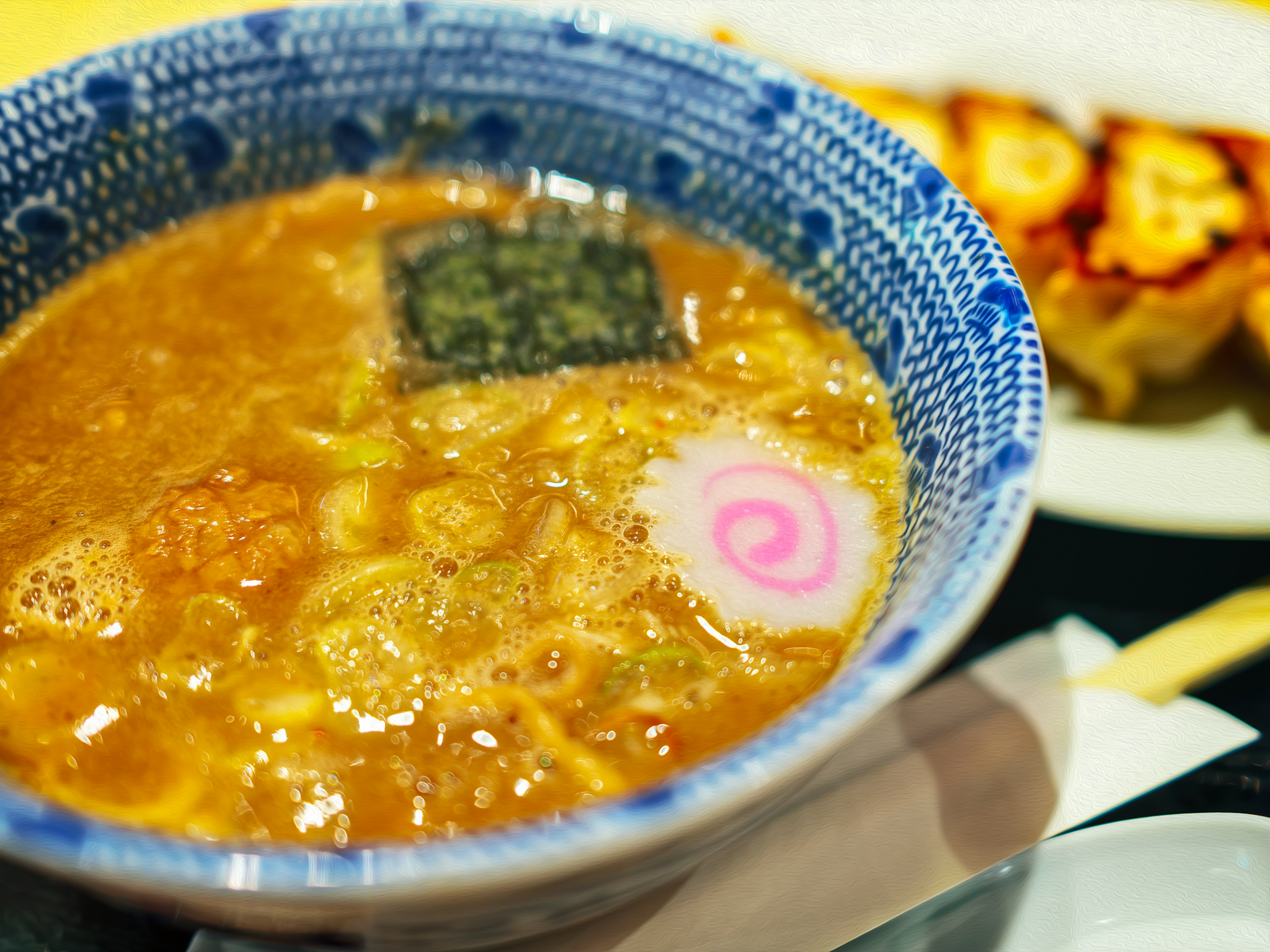 Ramen in einer blauen Schüssel mit Algen und Narutomaki-Garnituren neben gegrillten Gyoza