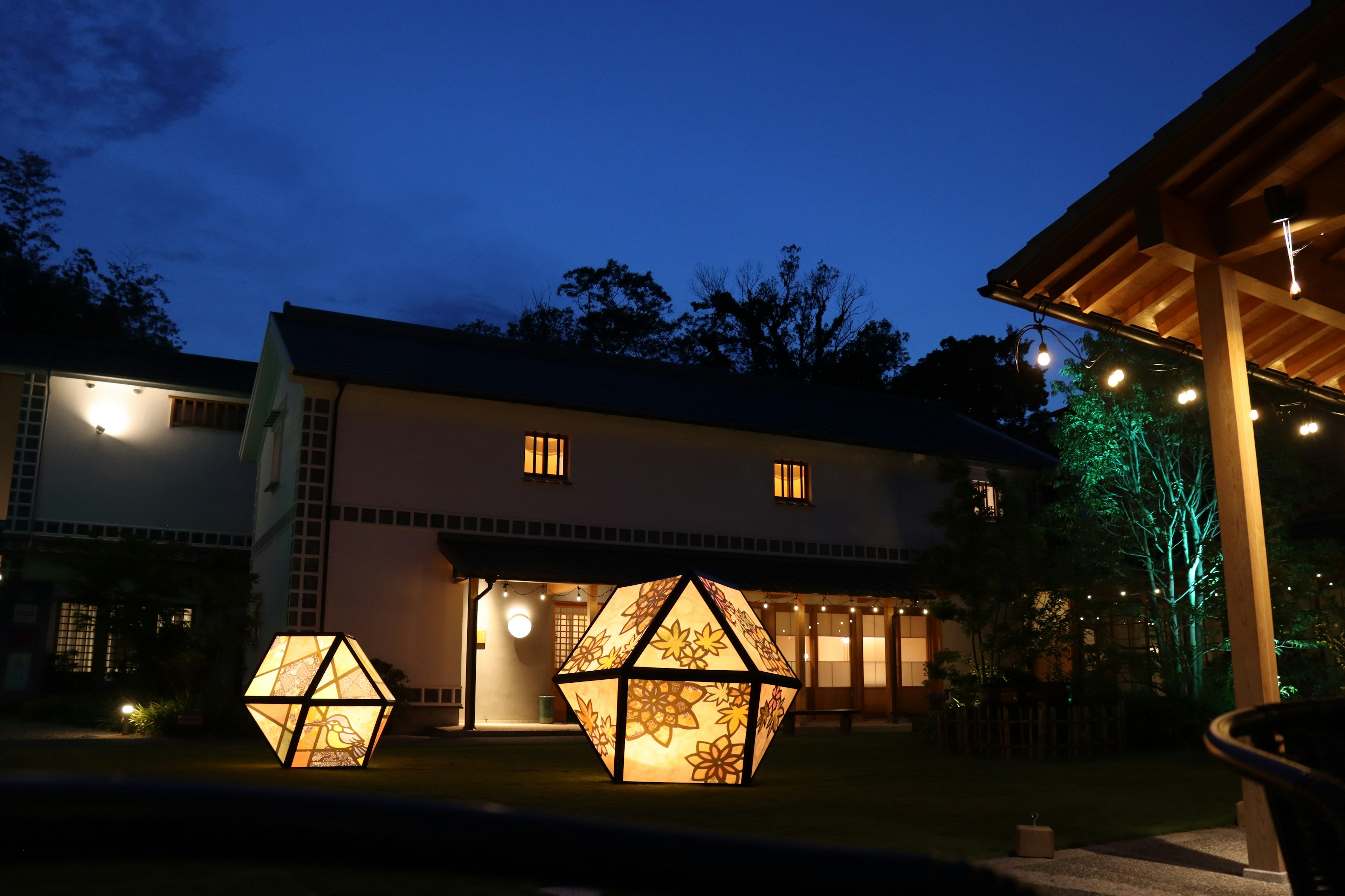 Lámparas geométricas iluminadas en un jardín por la noche con una casa al fondo