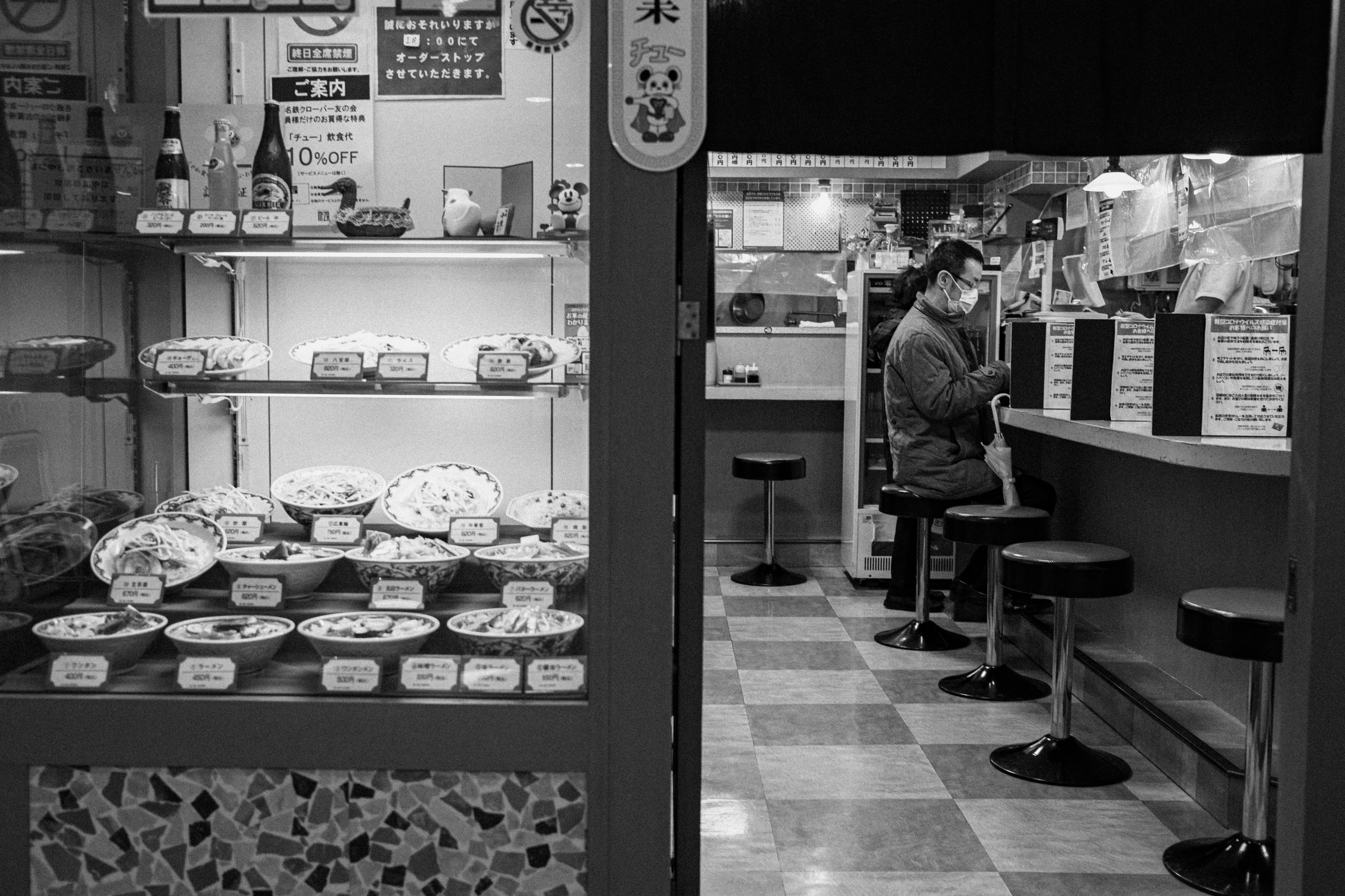 Foto en blanco y negro de un restaurante interior y exterior Exhibición de muestras de comida en la ventana y un cliente cenando en el mostrador