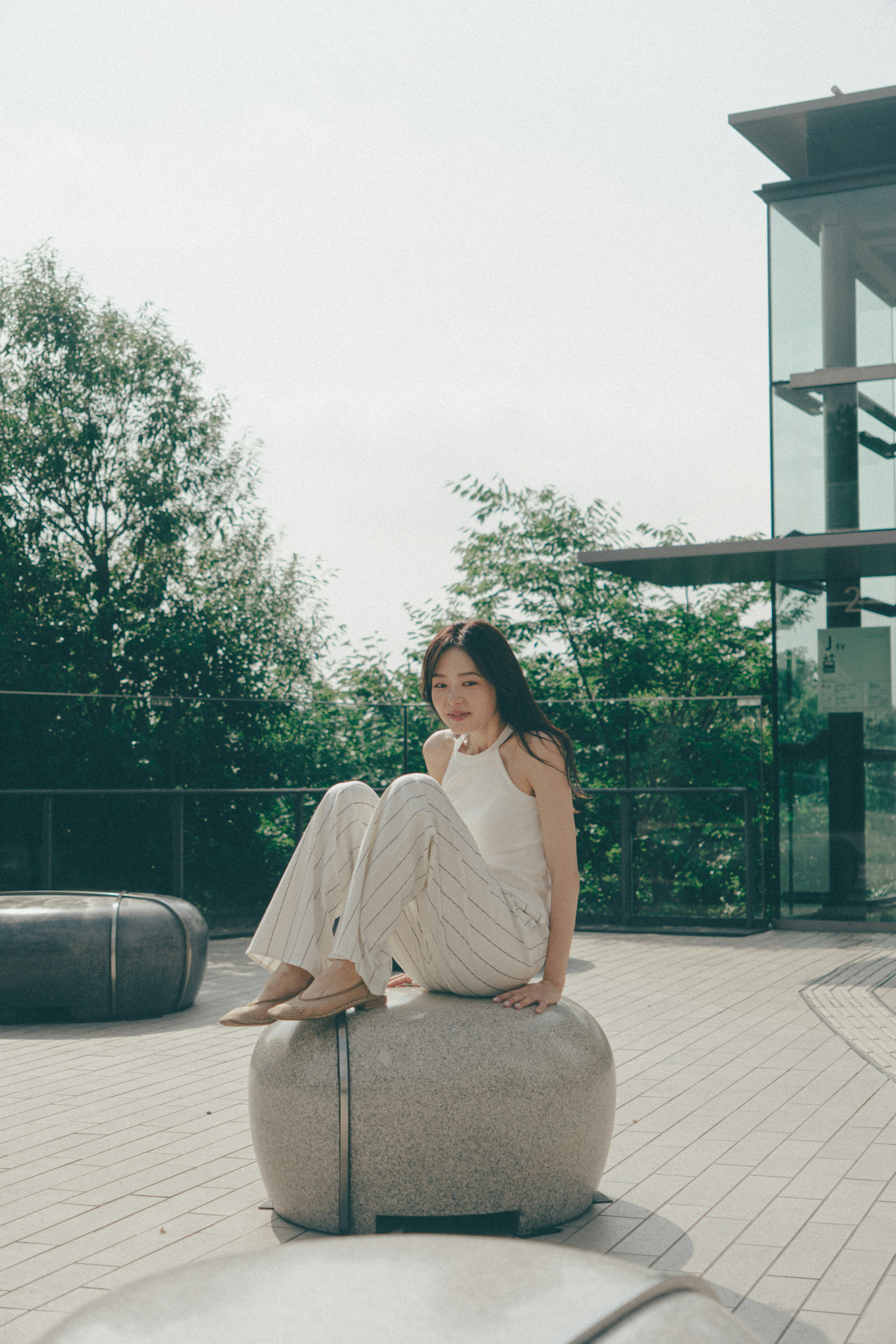 Woman sitting on a stone-like object outdoors with greenery and modern architecture in the background