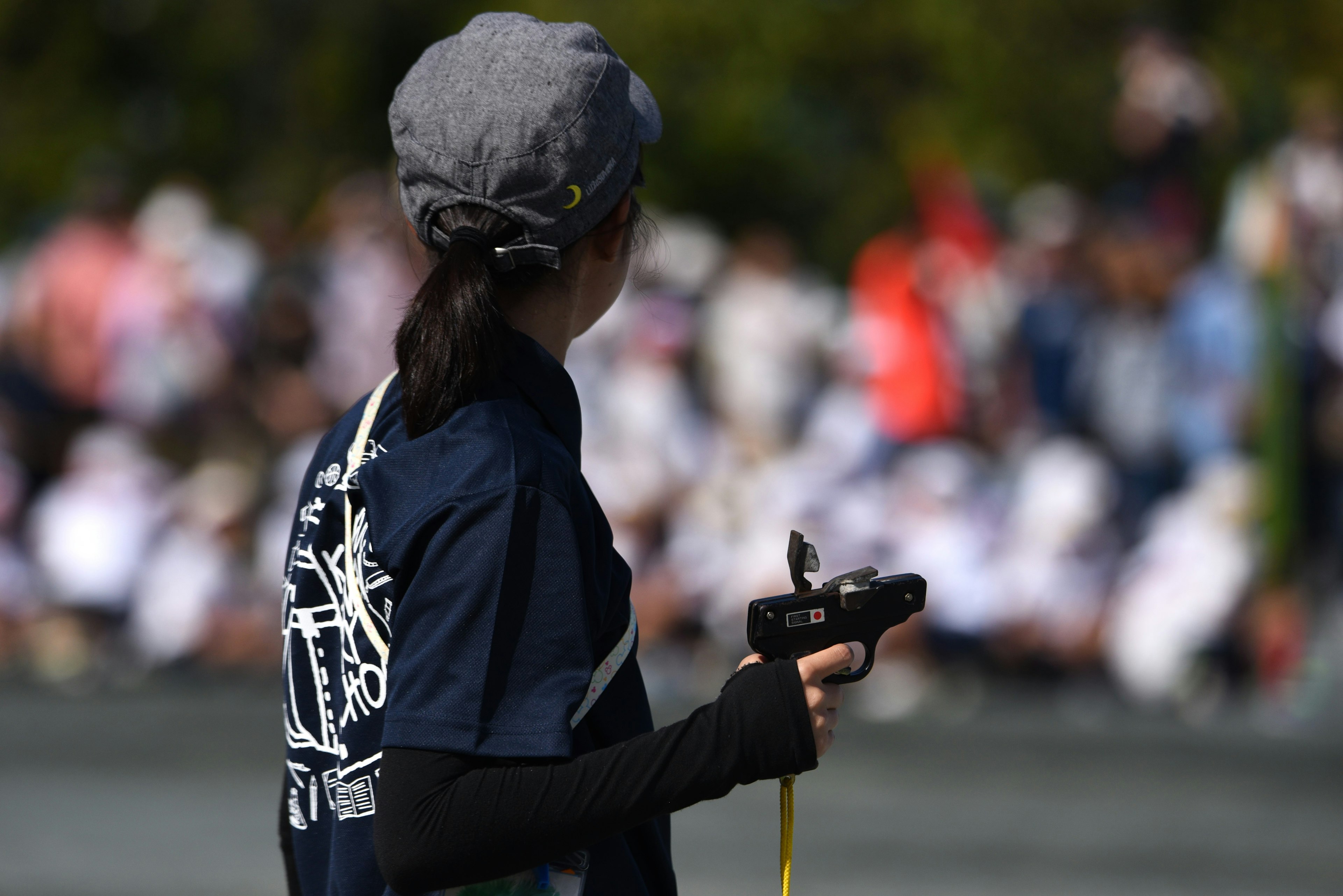 Frau beobachtet ein Sportereignis, trägt eine graue Mütze und schwarze Handschuhe, Design auf der Rückseite ihres Shirts, Zuschauer im Hintergrund