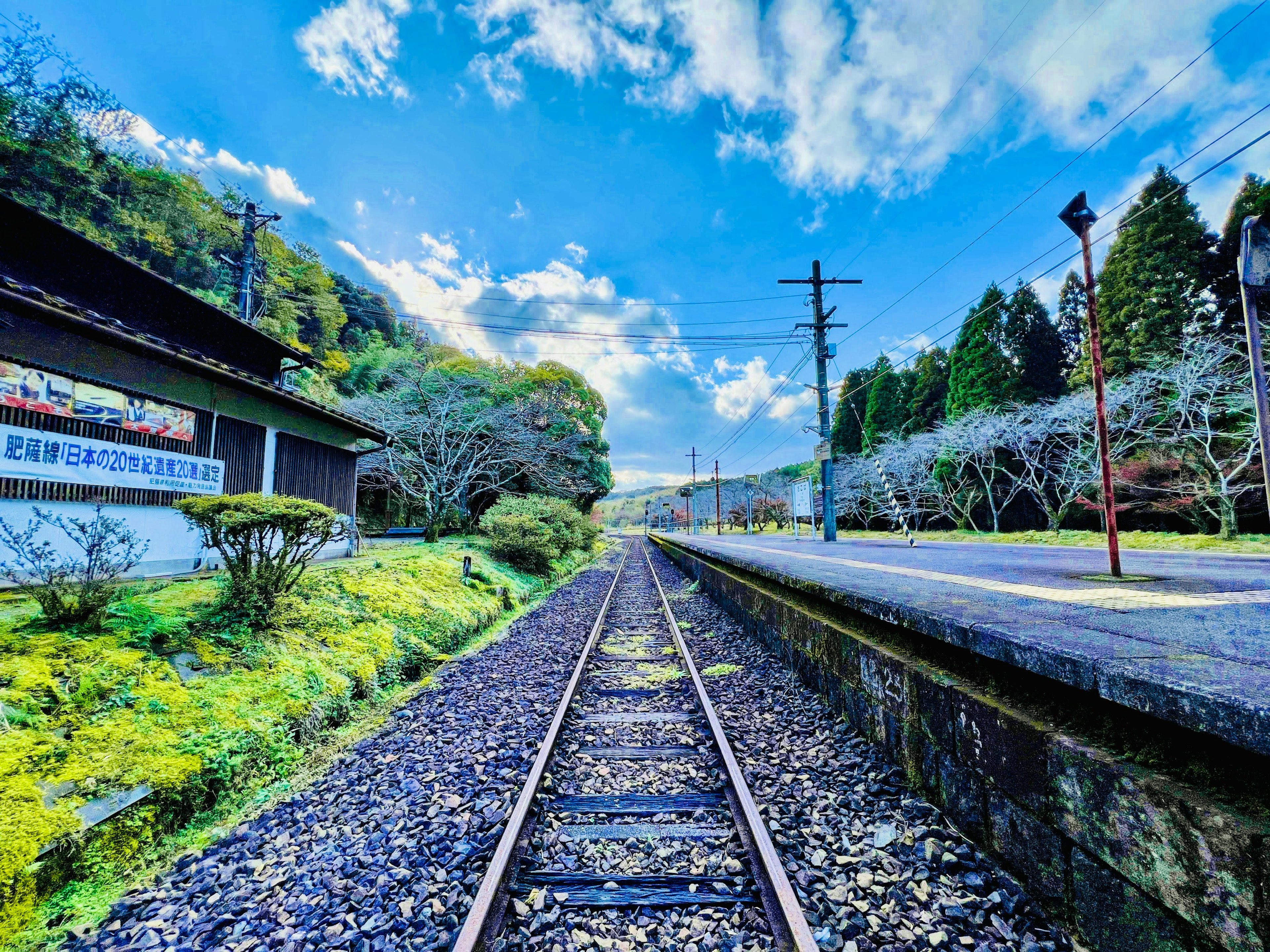 Jalur kereta yang tenang dikelilingi oleh pepohonan subur dan langit biru yang cerah