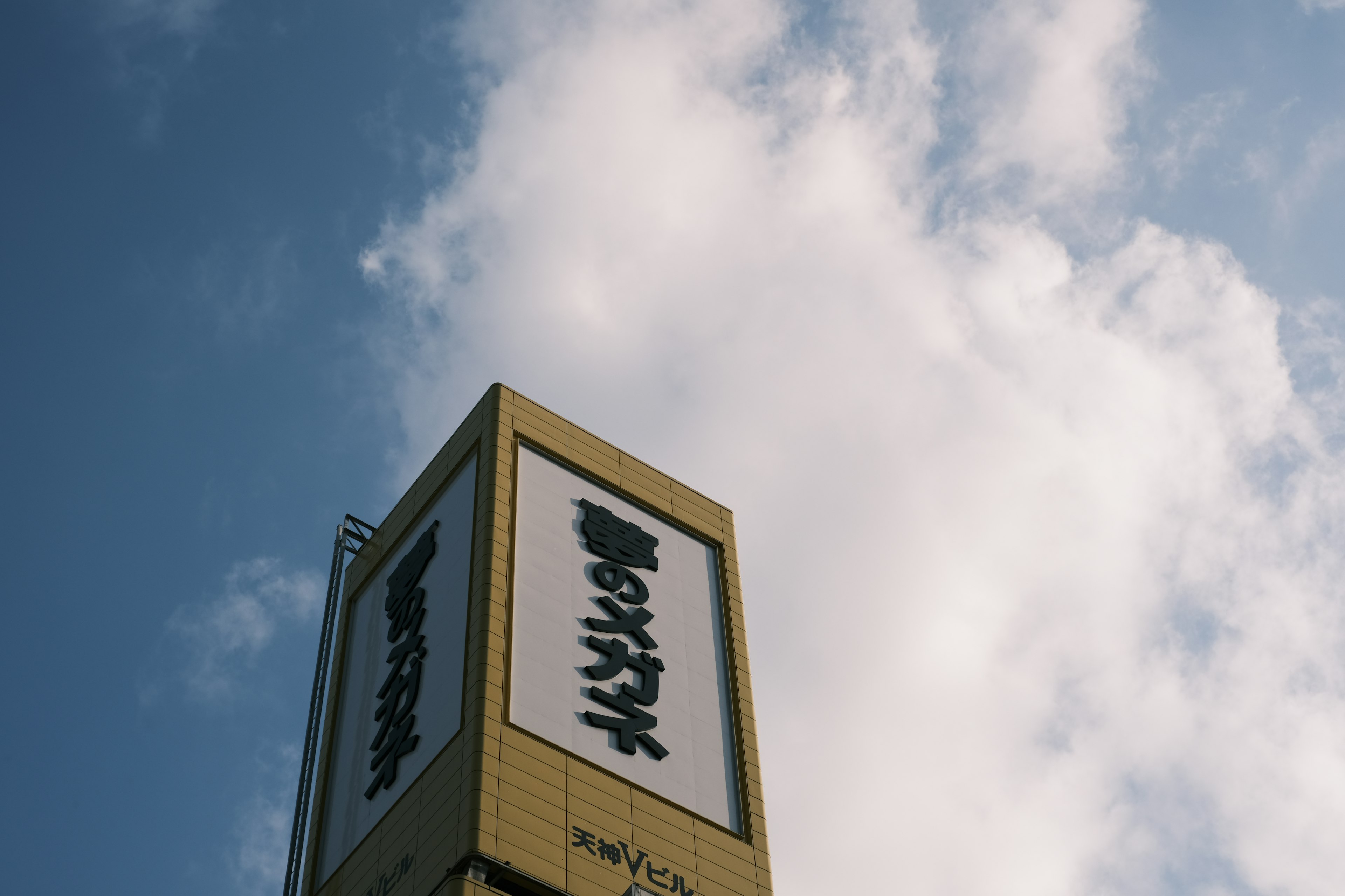 Cartel amarillo con texto en japonés contra un fondo de nubes blancas y cielo azul