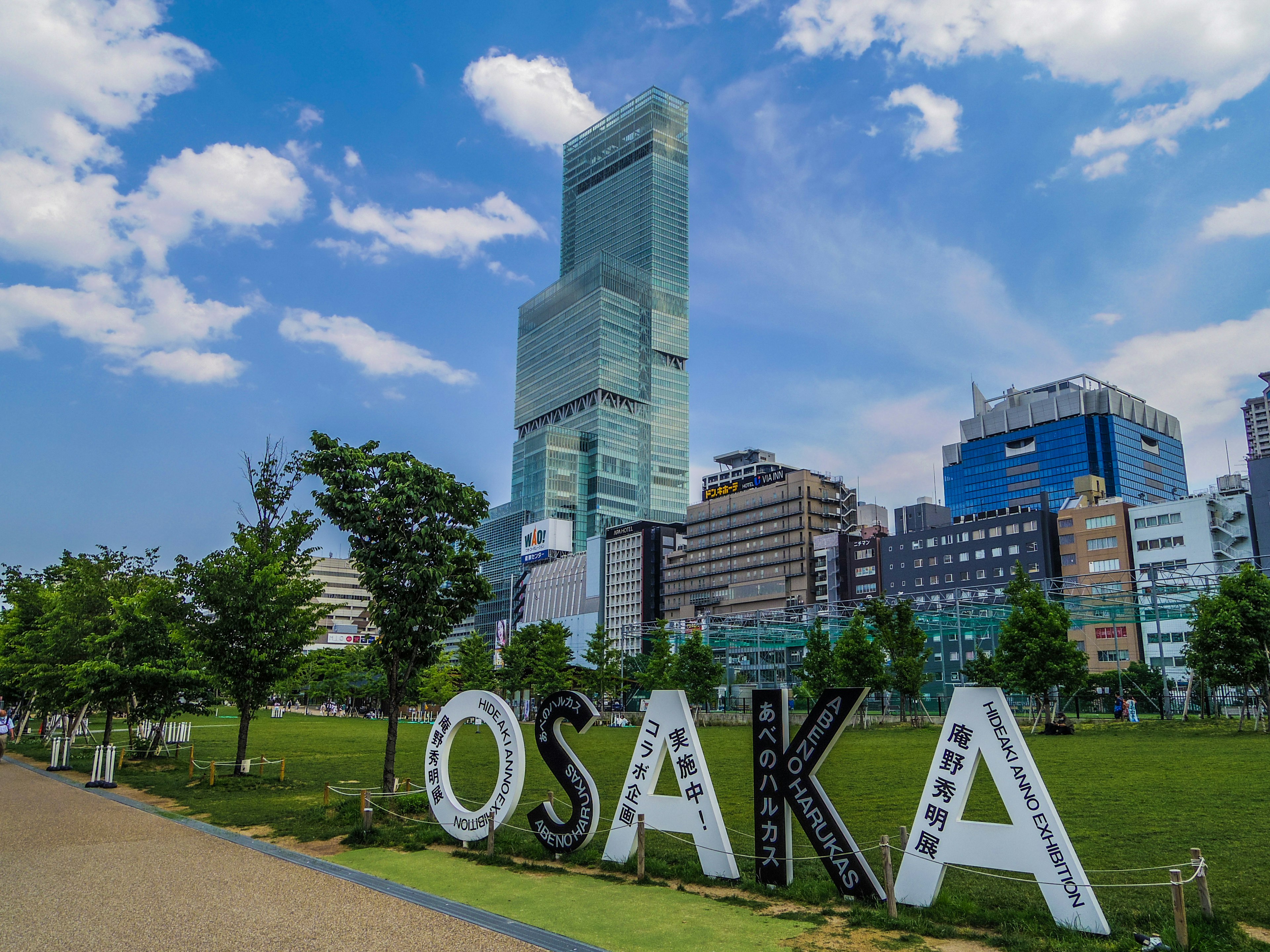 Großes Schild mit der Aufschrift Osaka in einem Park mit Wolkenkratzern und blauem Himmel