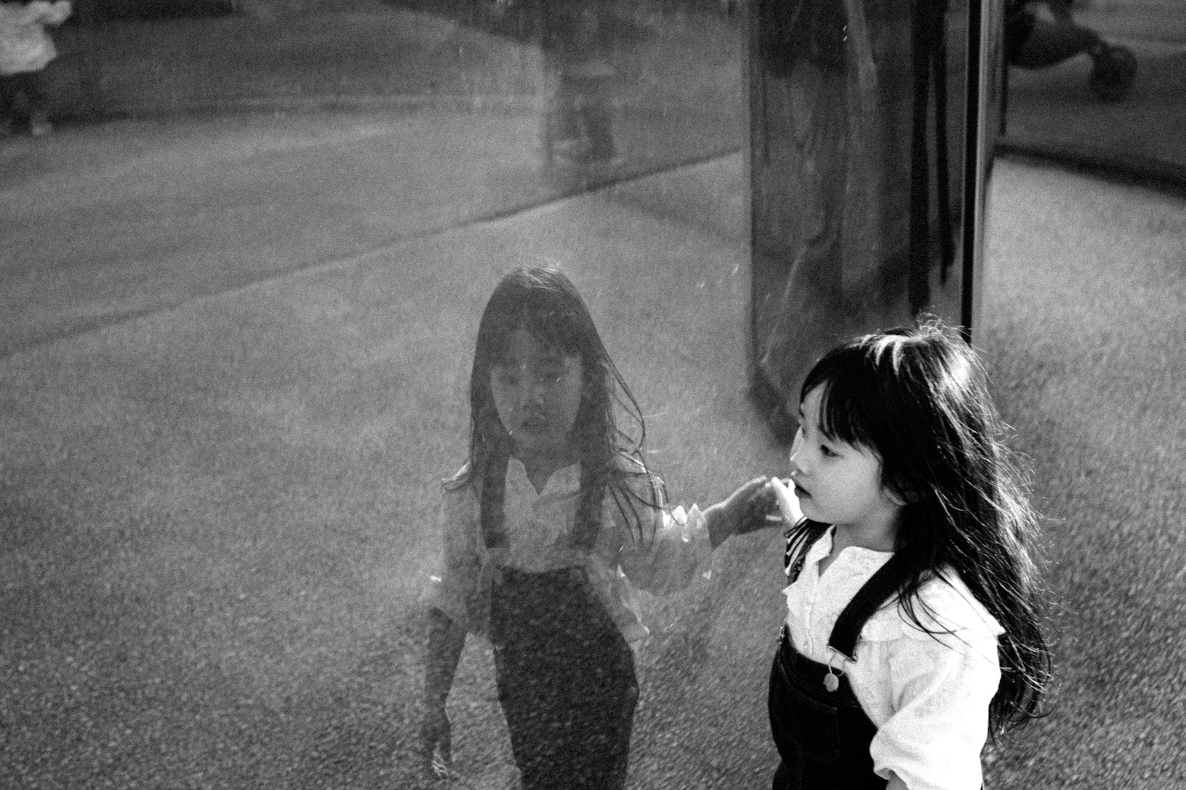 Black and white photo of a girl standing in front of a mirror her reflection visible in the glass