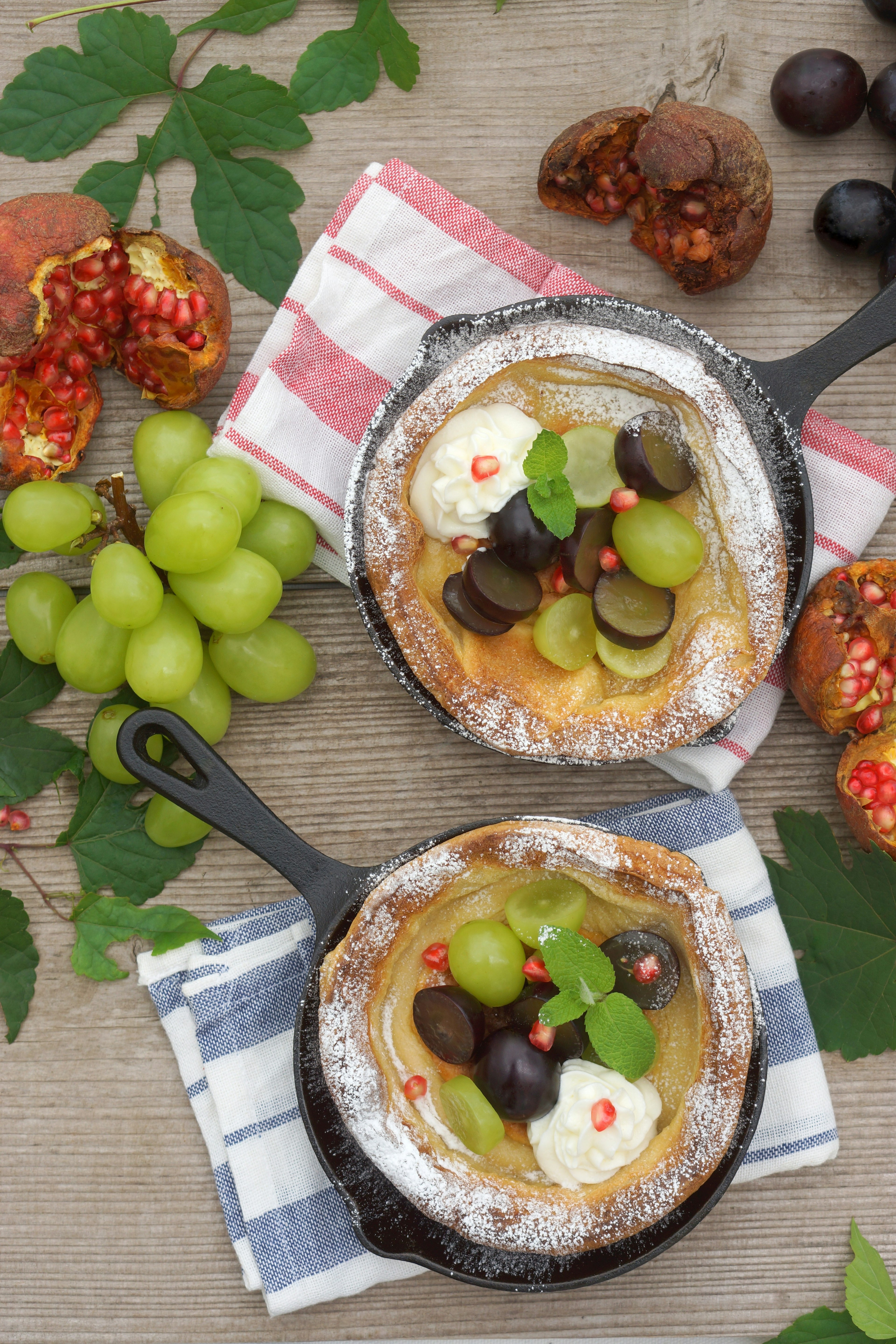 Zwei Pfannen mit Obst- und Cremedessert umgeben von Trauben und Granatapfel