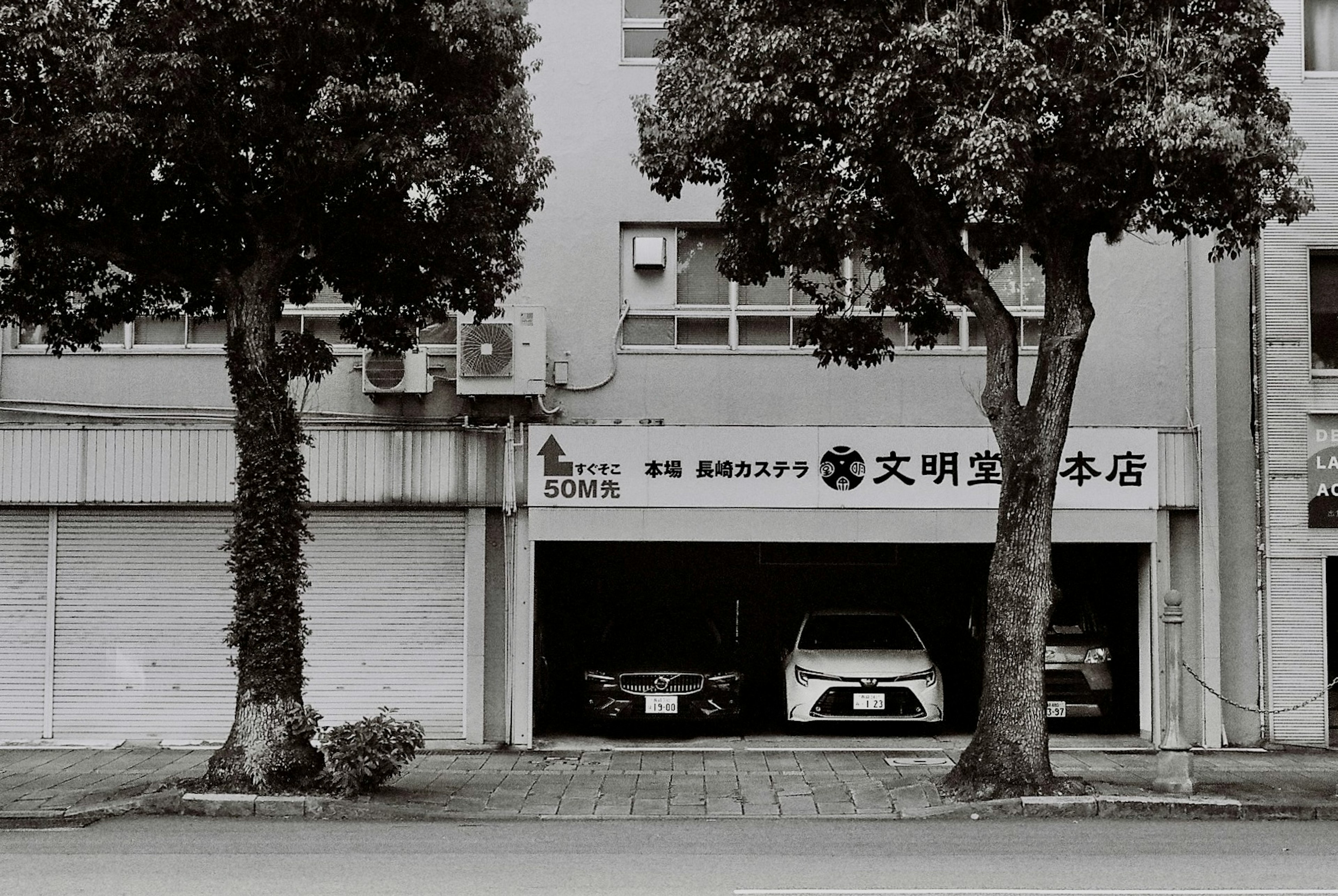 白黒の写真の中に駐車場があり、二台の車が見える。木が側に立っており、建物の看板には日本語の文字がある。