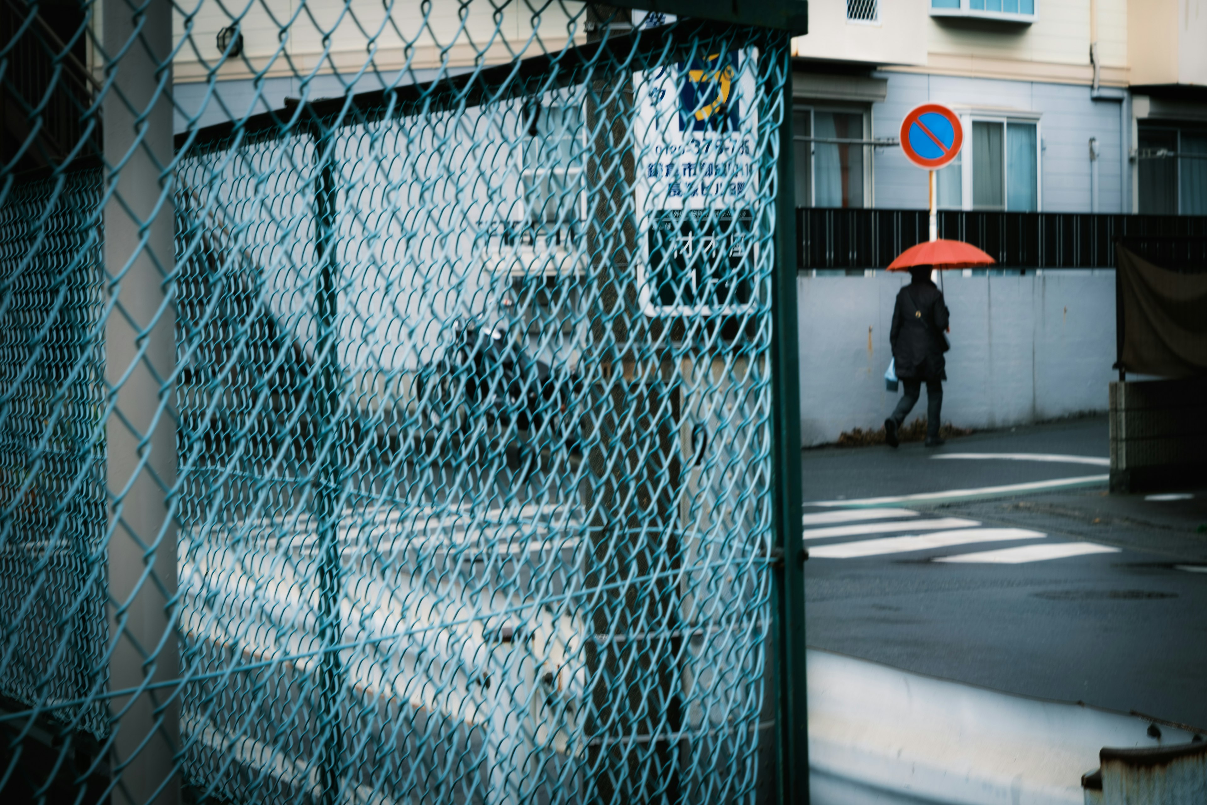 Eine Person mit einem roten Regenschirm, die durch einen Maschendrahtzaun mit Gebäuden im Hintergrund gesehen wird