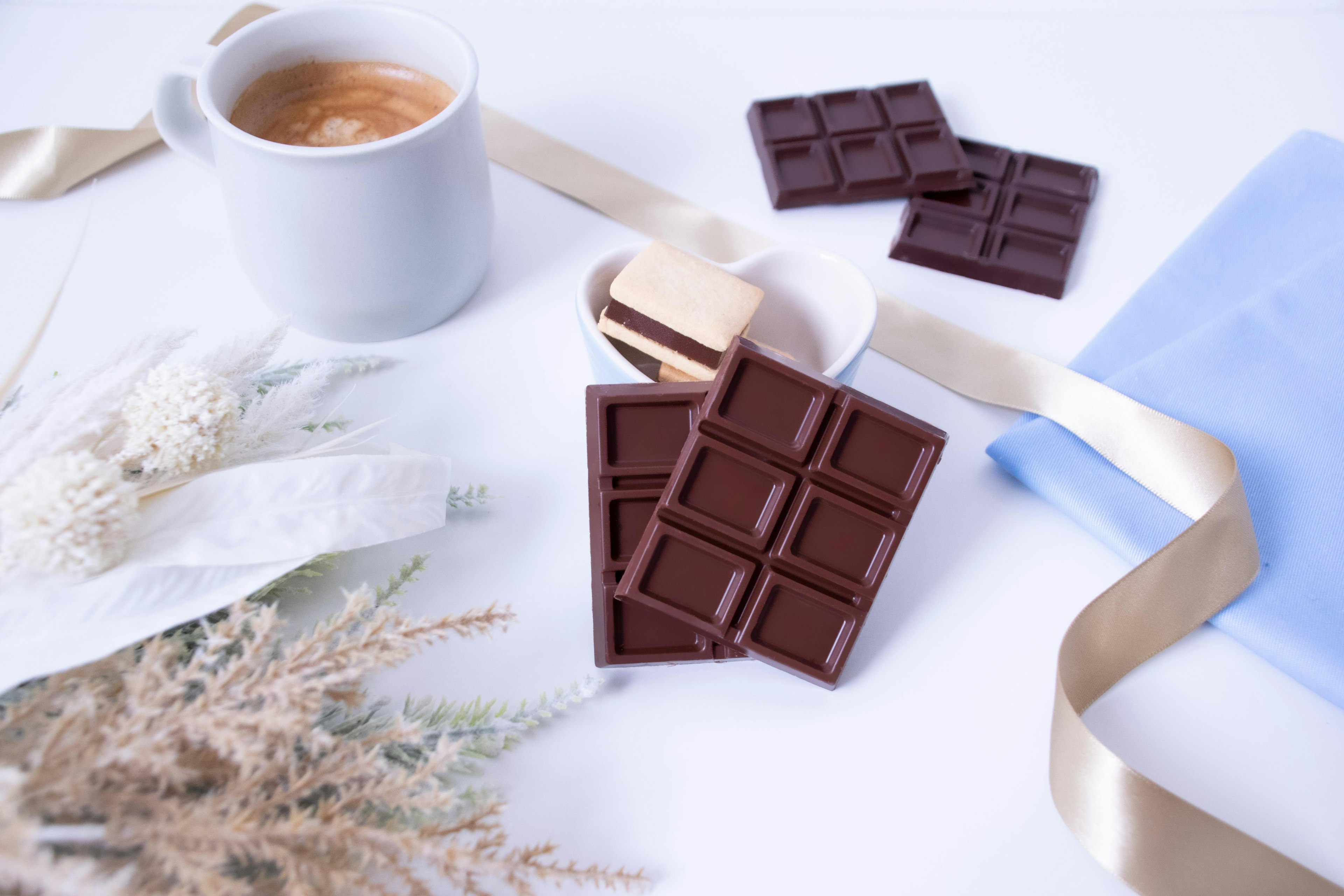 A cup of coffee beside chocolate bars with a ribbon and dried flowers on a table