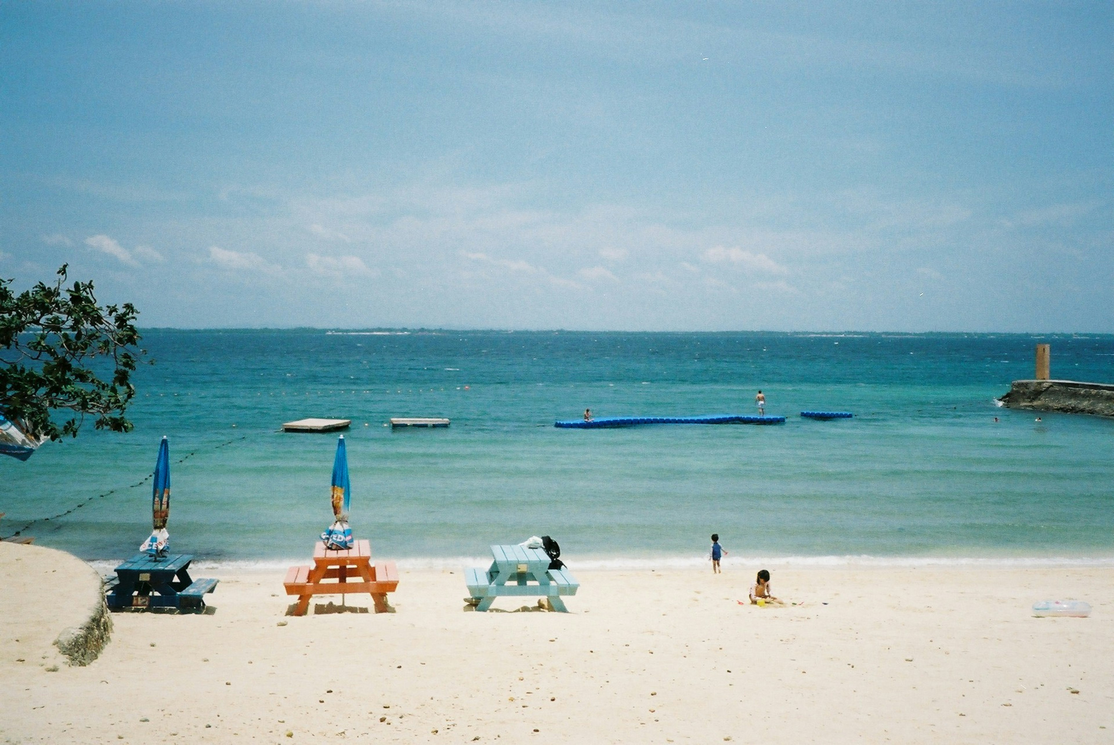 Bunte Picknicktische an einem Sandstrand mit klarem blauem Meer und Booten in der Ferne