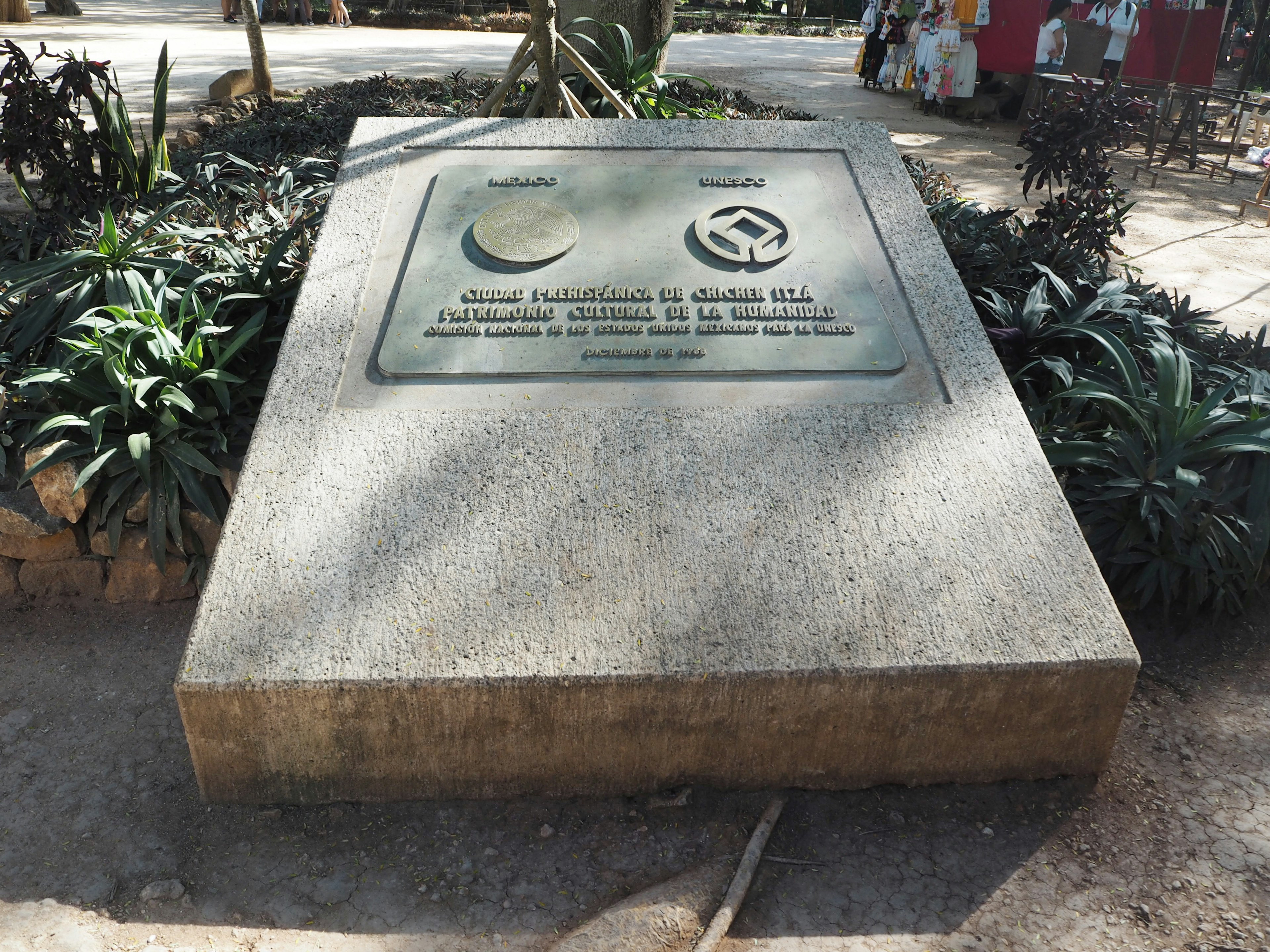 Monumen batu di taman dengan plakat peringatan dikelilingi tanaman