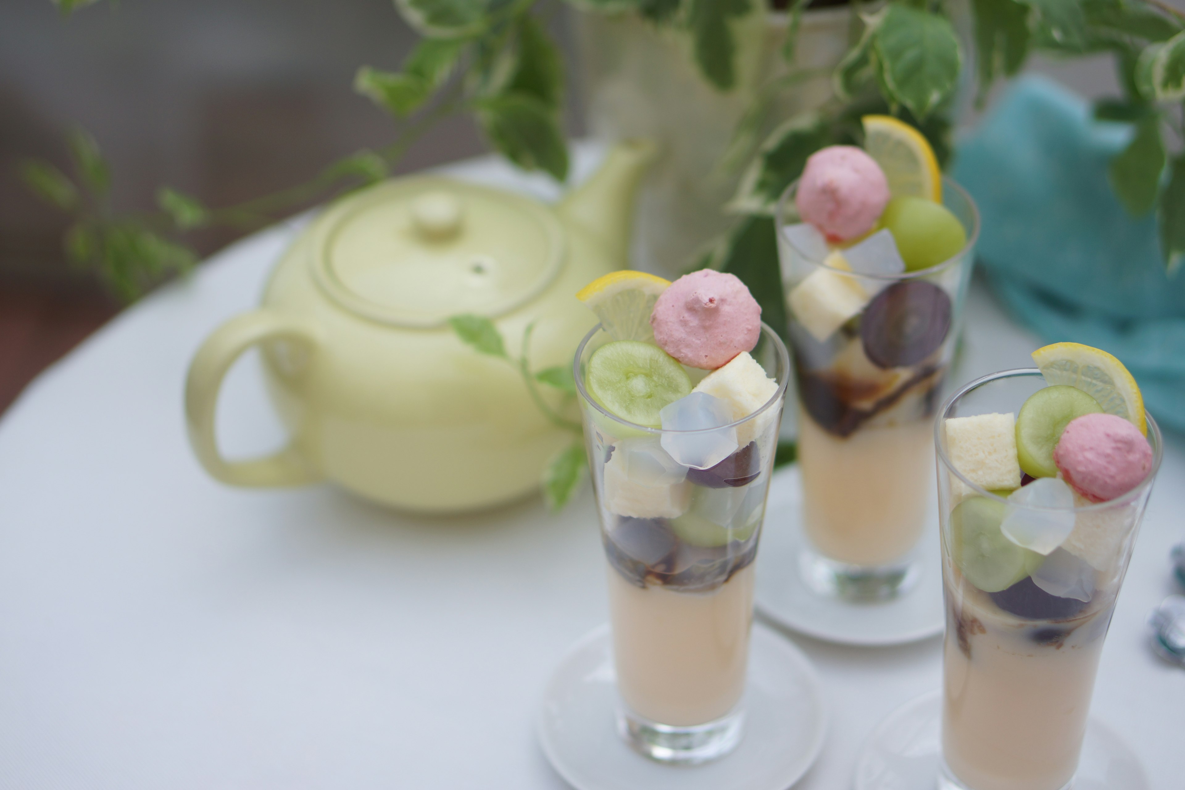 Trois boissons dessert colorées dans des verres avec une théière jaune et des plantes vertes en arrière-plan