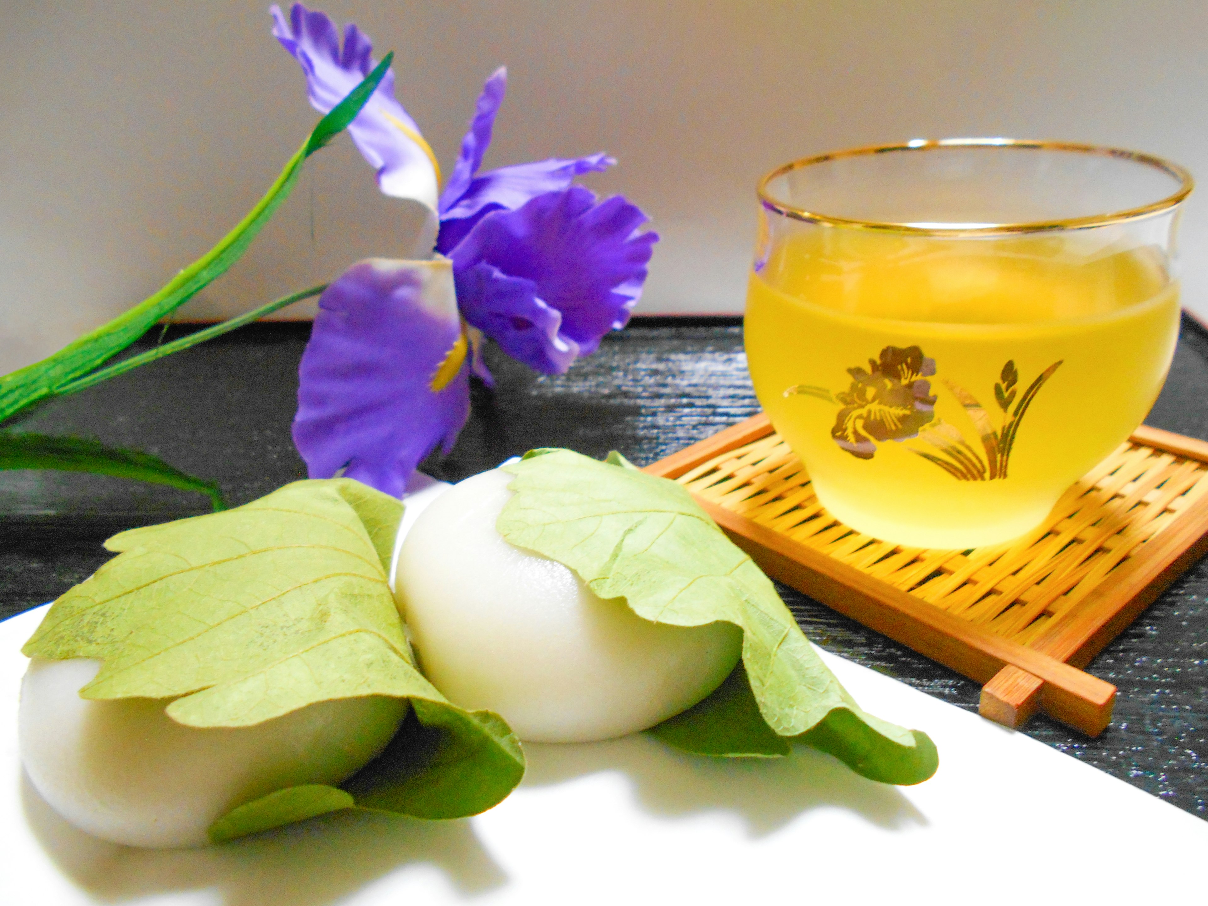 A serene table setting featuring white Japanese sweets wrapped in green leaves with a cup of tea and a purple flower