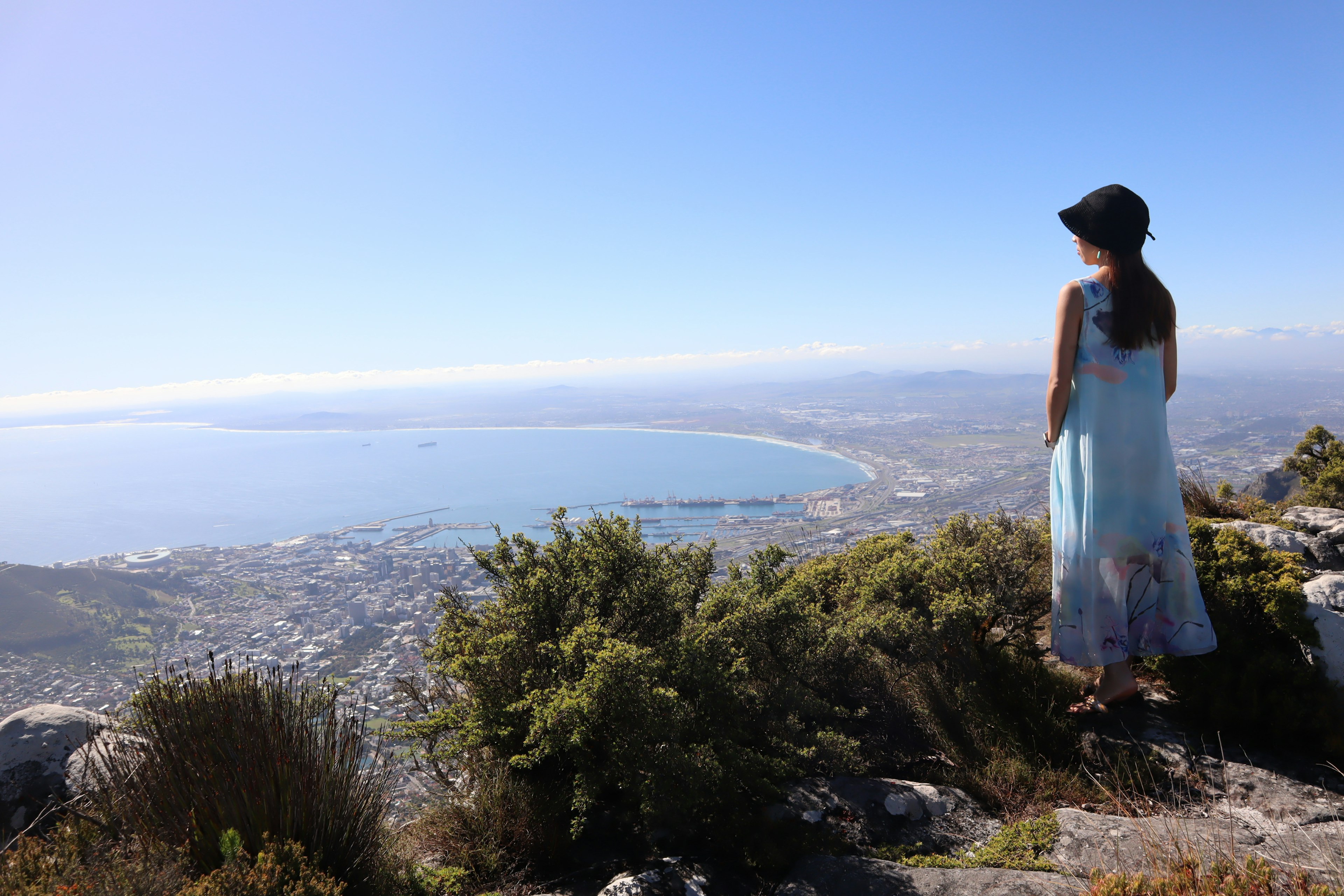 Frau in blauem Kleid, die von einem Berggipfel auf die Küste blickt