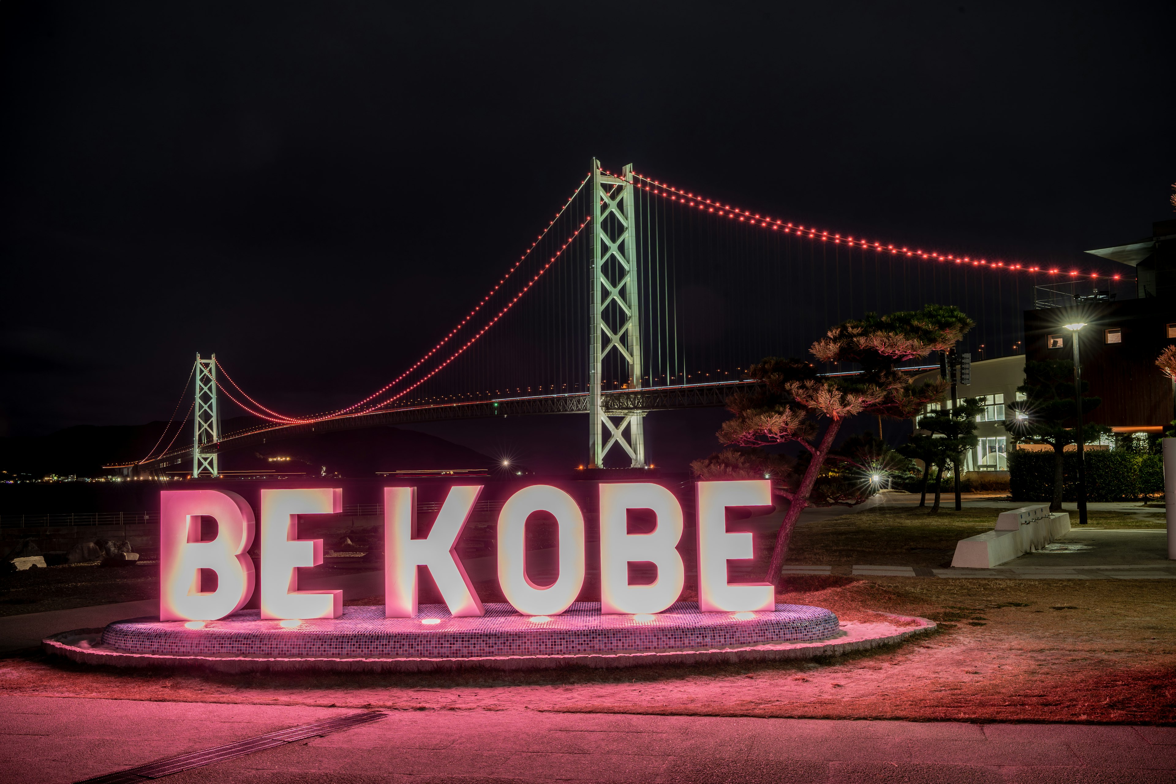 Large illuminated letters spelling BE KOBE with a decorative bridge in the background at night