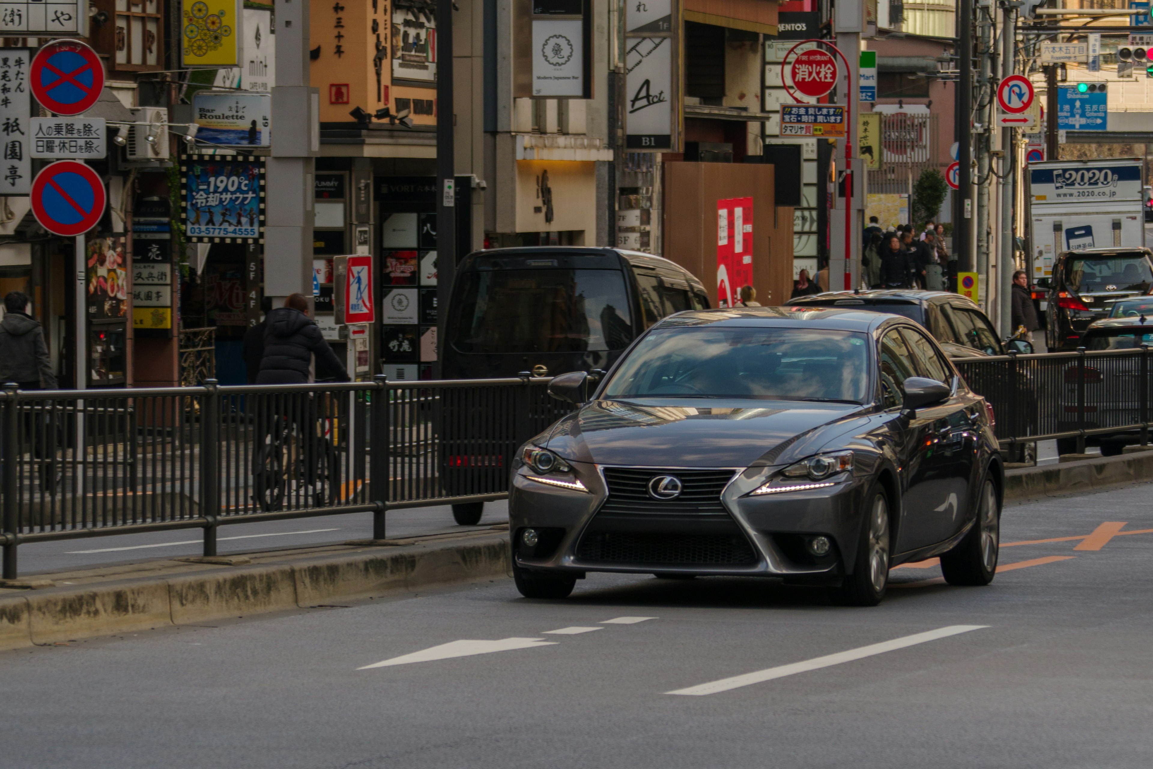 都市の通りを走る黒いレクサスの車周囲には人々と商店が並んでいる