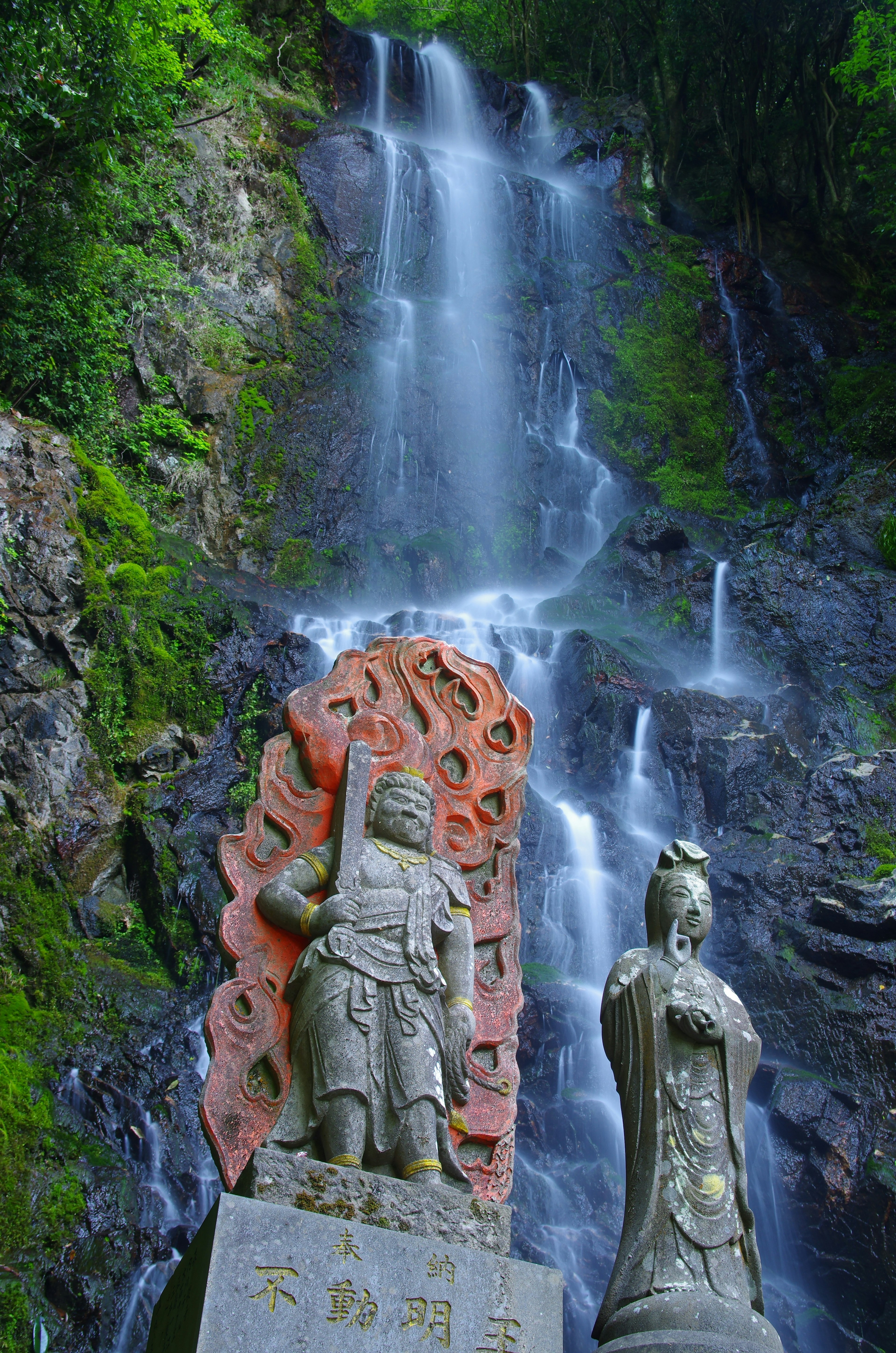 Zwei Steinstatuen vor einem Wasserfall Eine Statue stellt einen Krieger dar Die andere Statue zeigt eine Frau Umgeben von grünen Bergen