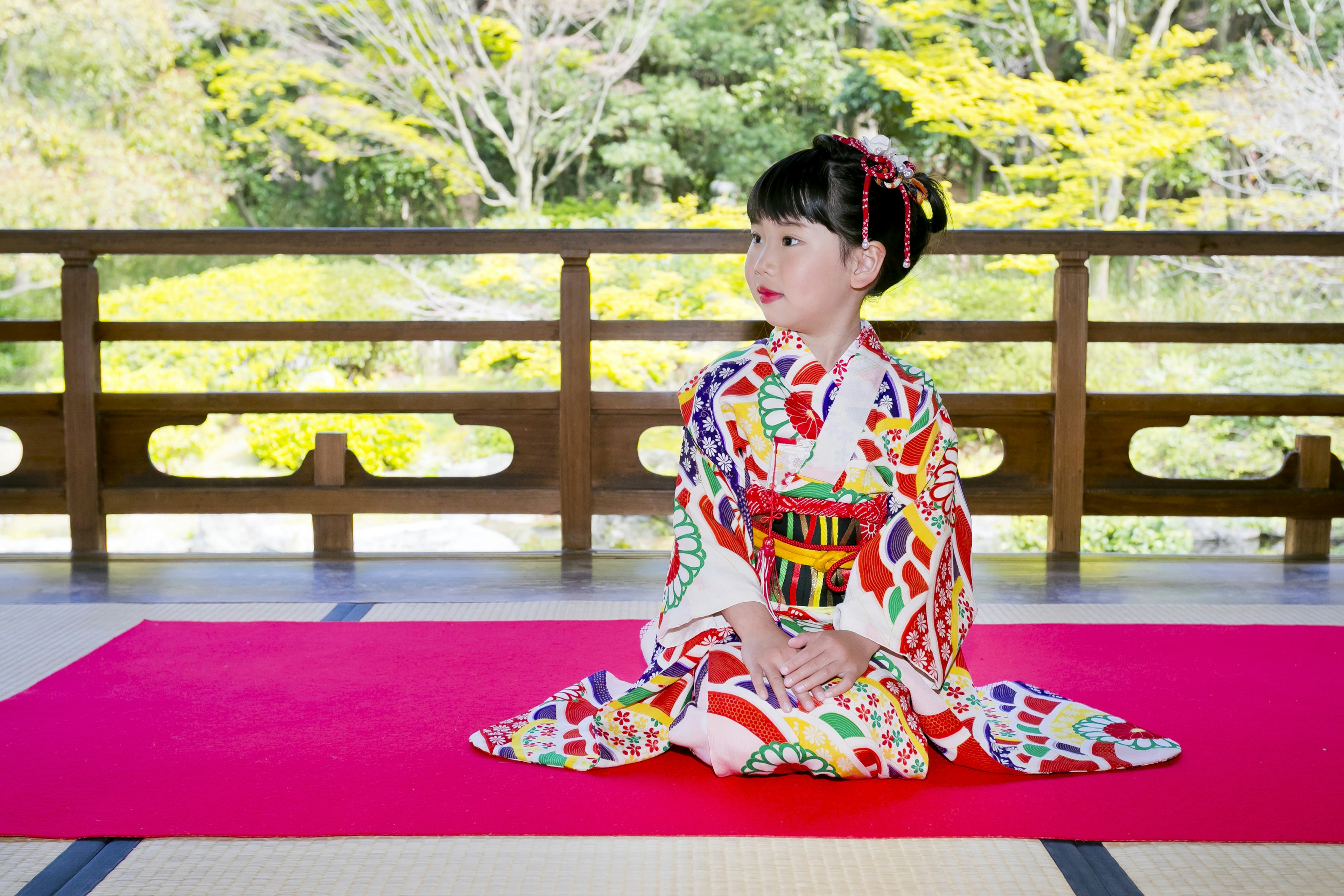 Gadis muda mengenakan kimono berwarna-warni duduk di atas permadani merah taman Jepang tradisional di latar belakang