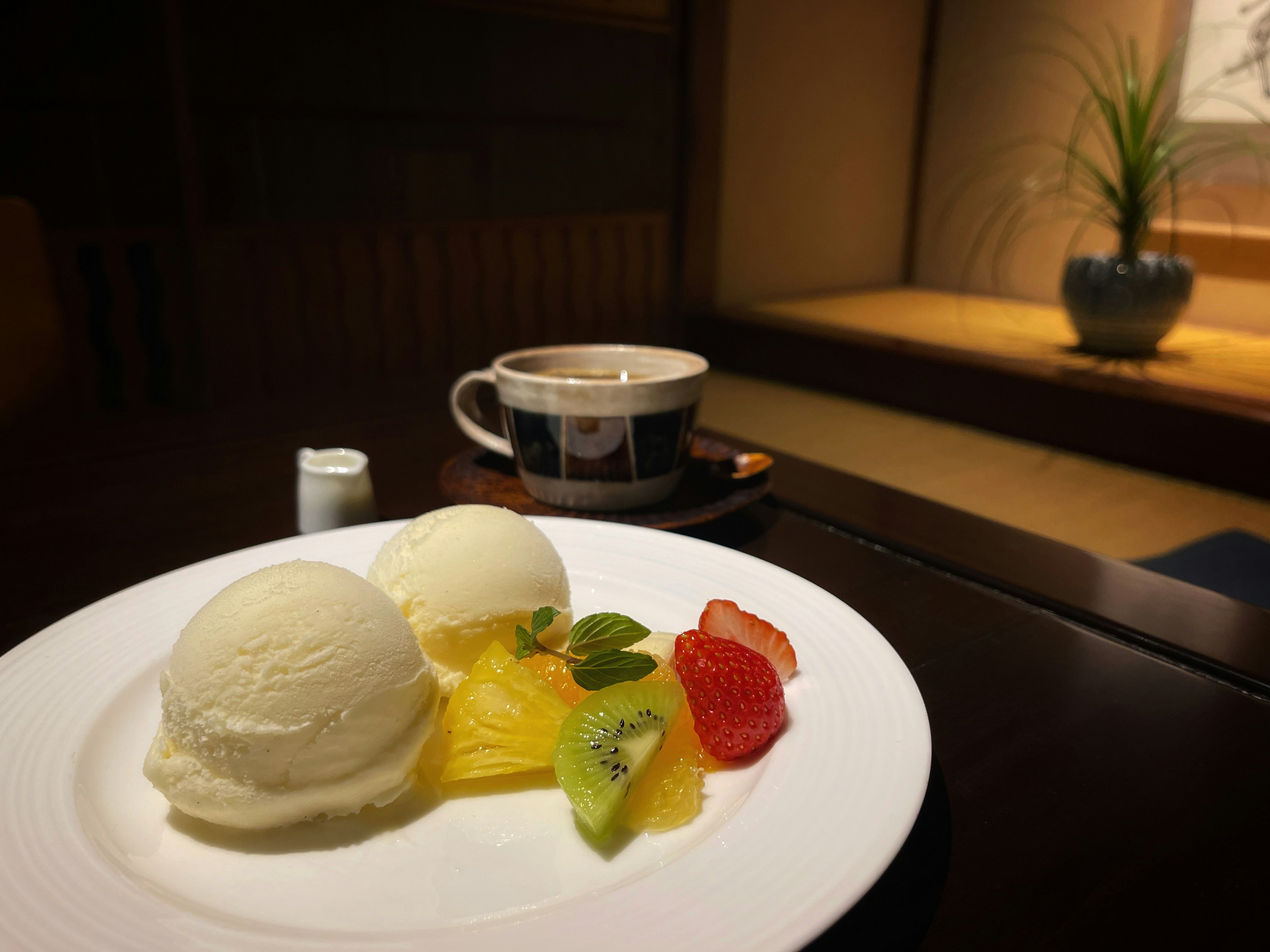 Un piatto con gelato bianco e un assortimento di frutta accanto a una tazza di caffè su un tavolo