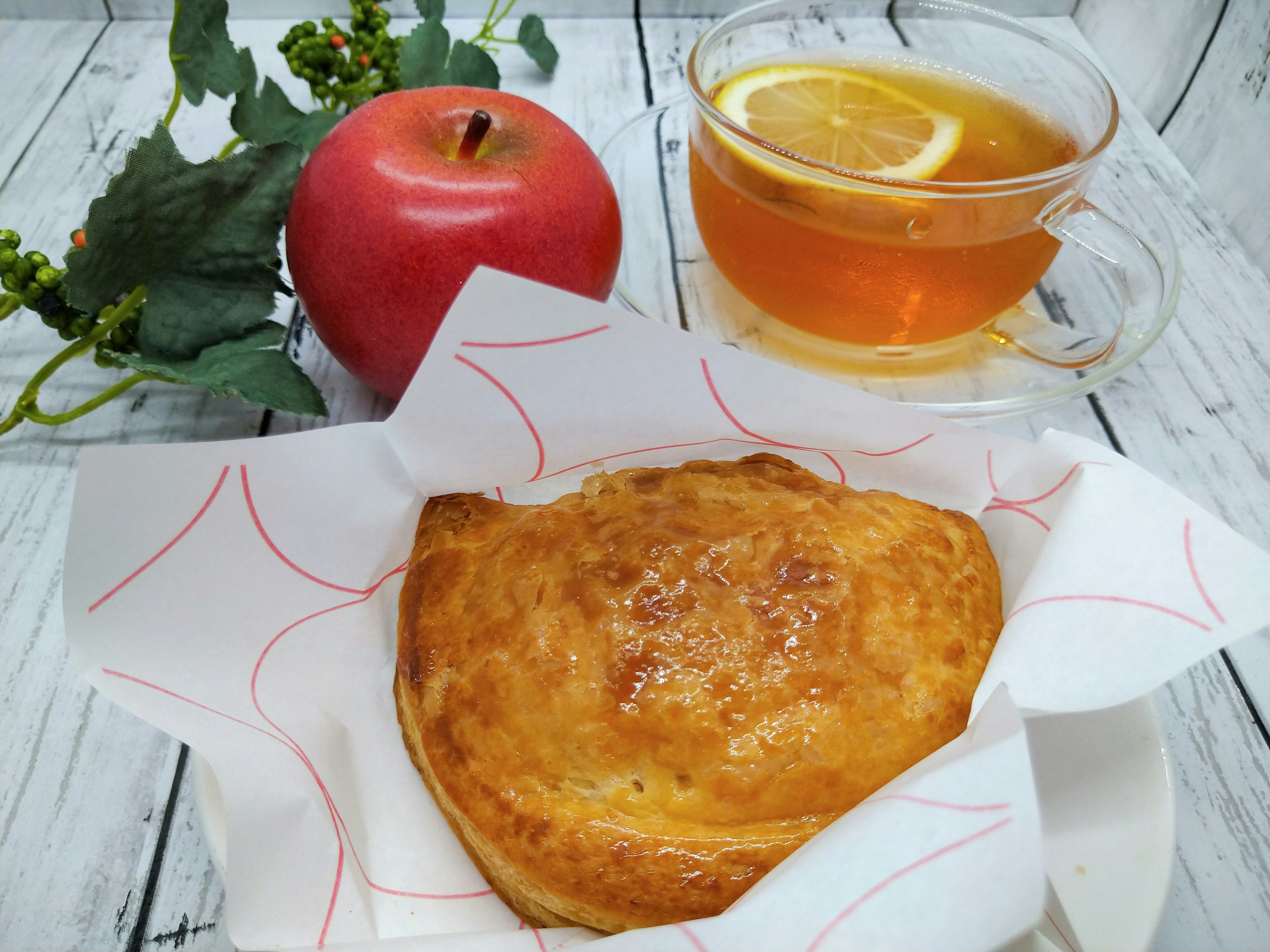 A golden pastry next to a red apple and a cup of tea with lemon slices