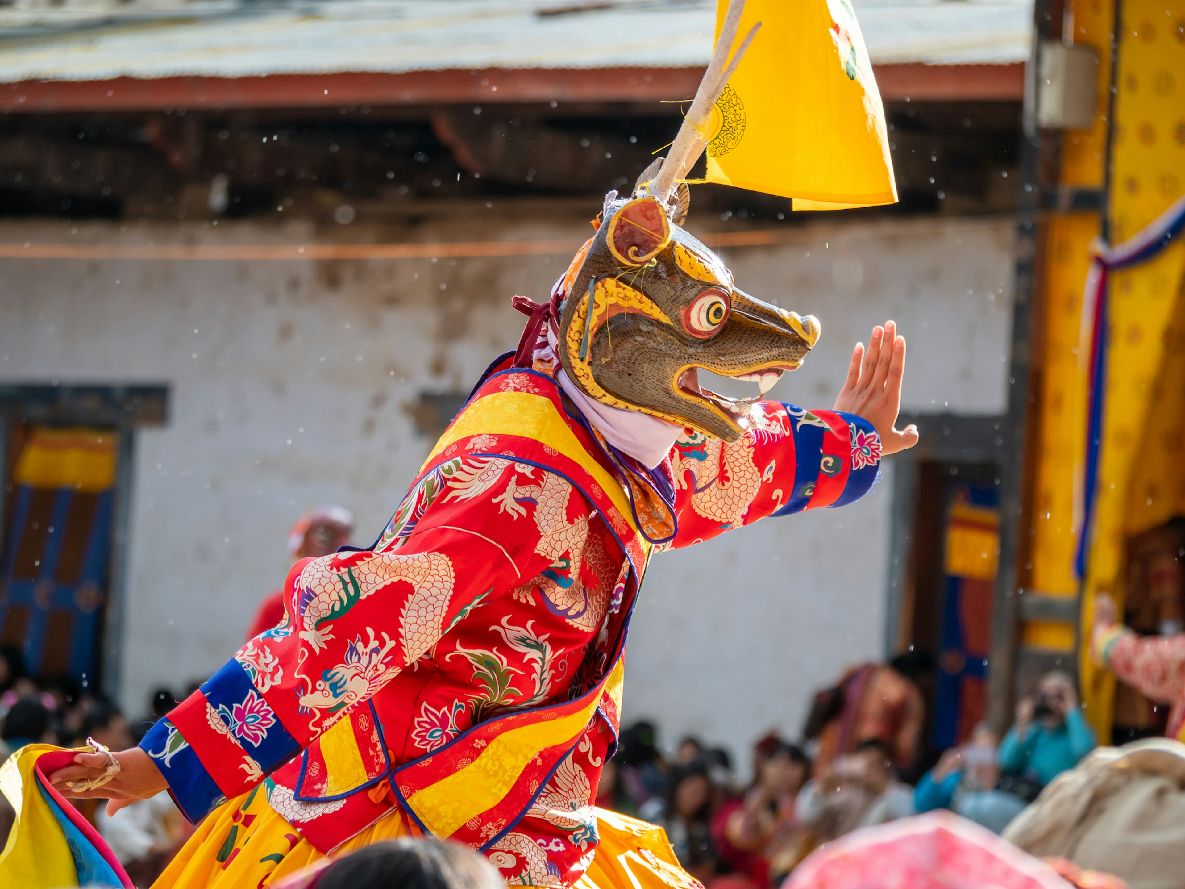 A dancer in a colorful costume wearing a mask raises one hand