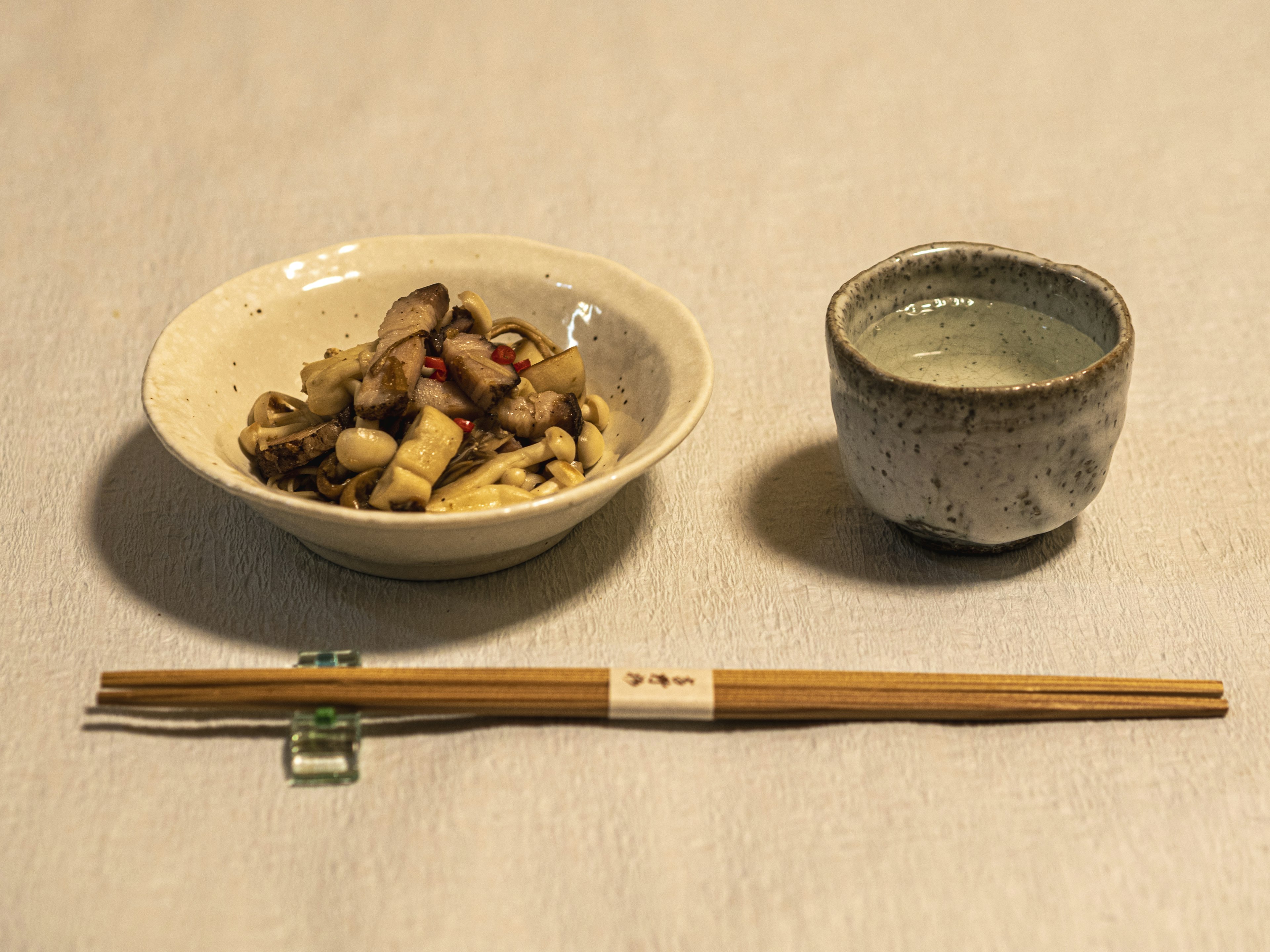 Un plato de champiñones mezclados en un tazón blanco junto a una taza de sake de cerámica y palillos de madera