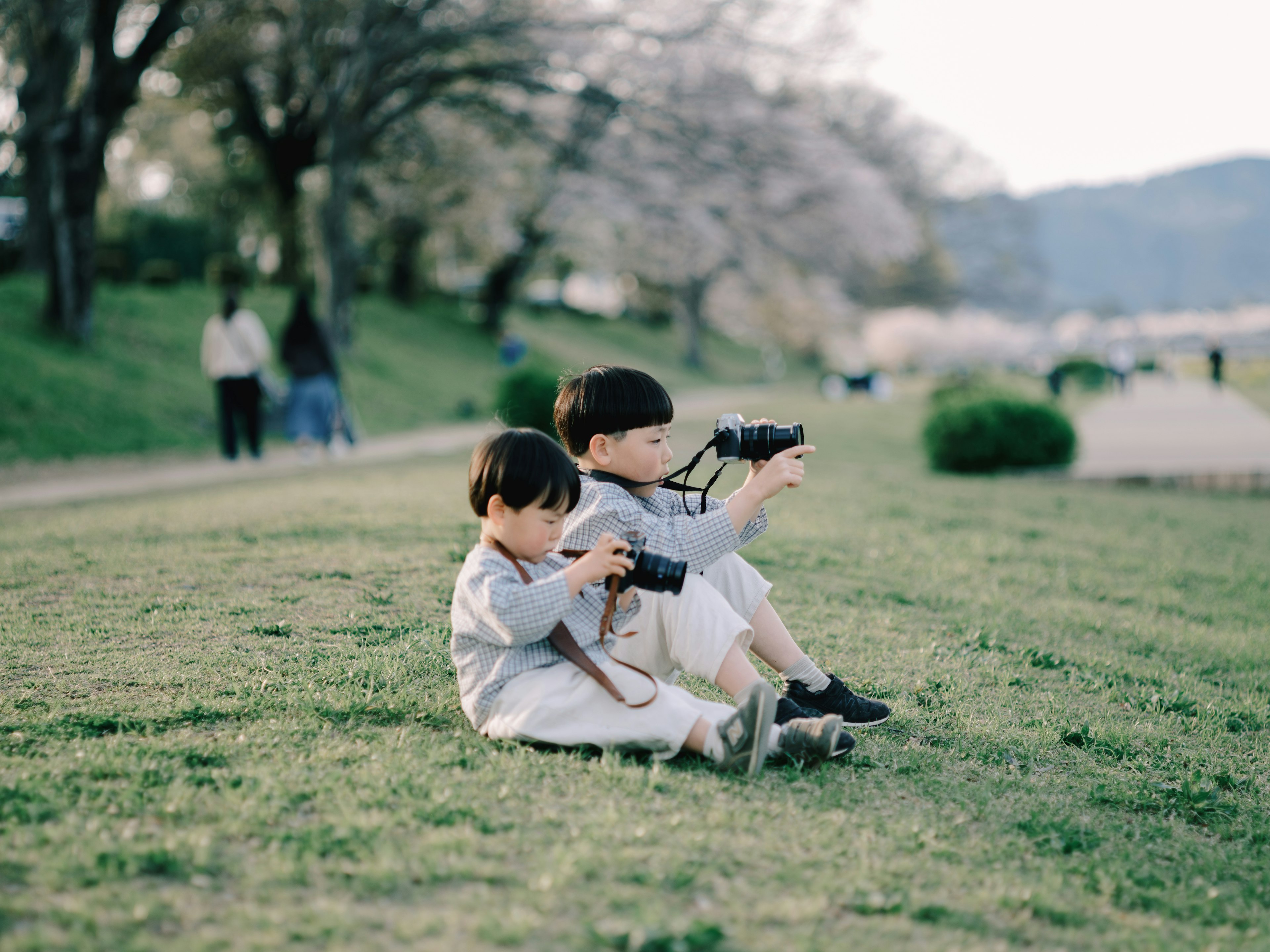 Two children sitting on grass in a park holding cameras