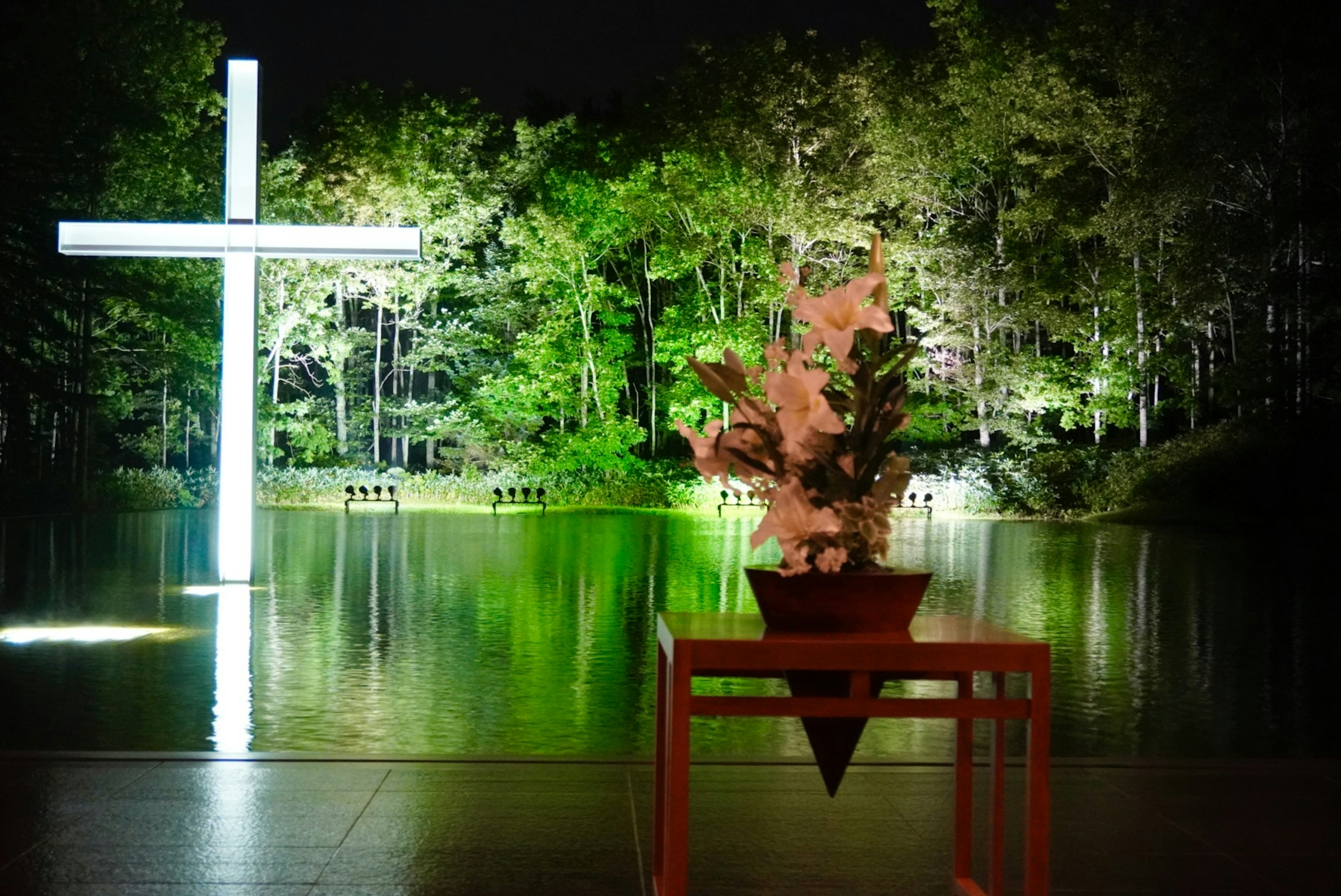 Illuminated cross beside a serene lake surrounded by lush greenery and a floral arrangement in the foreground