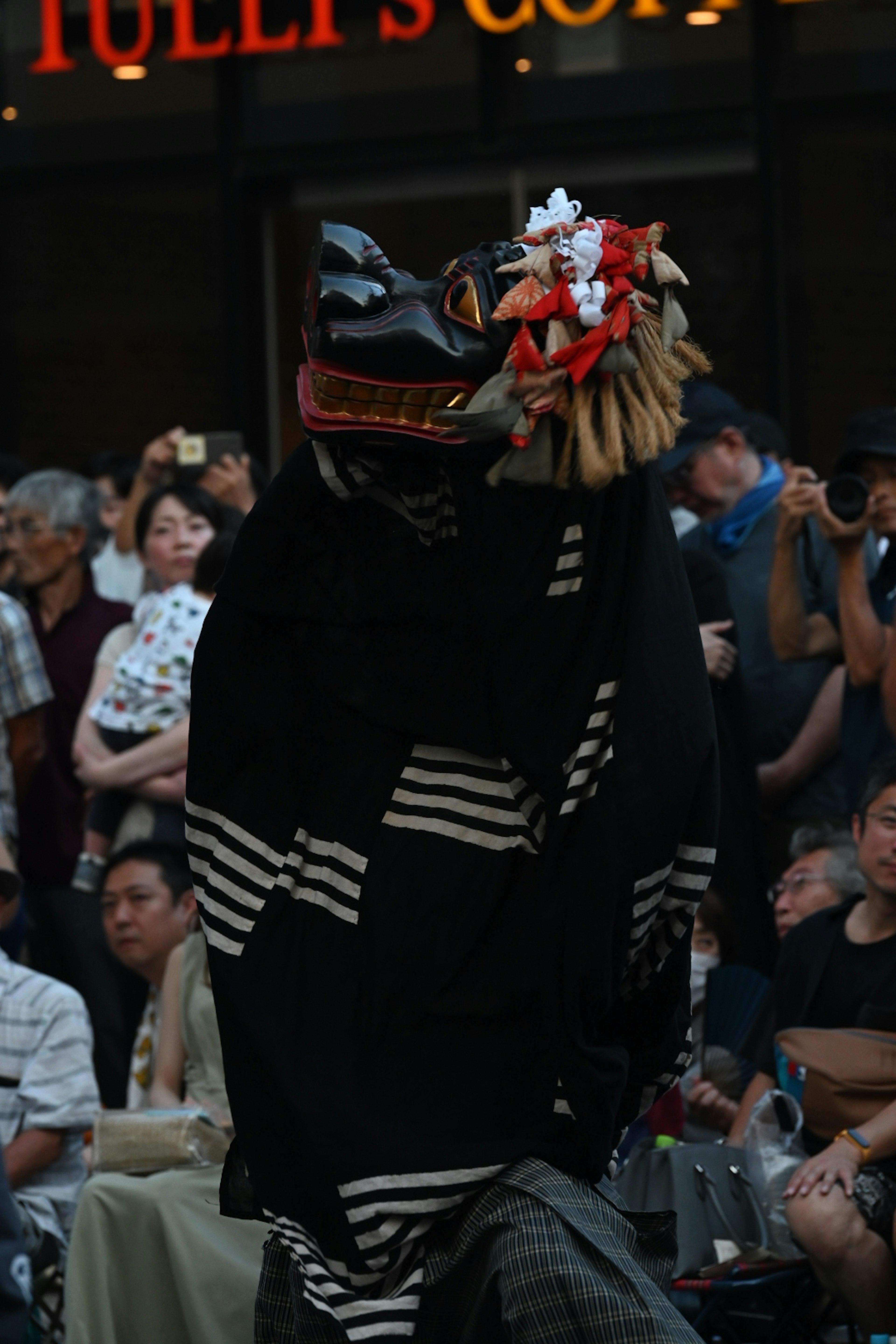 A performer in traditional lion dance attire with a black costume and floral decorations engaging an audience