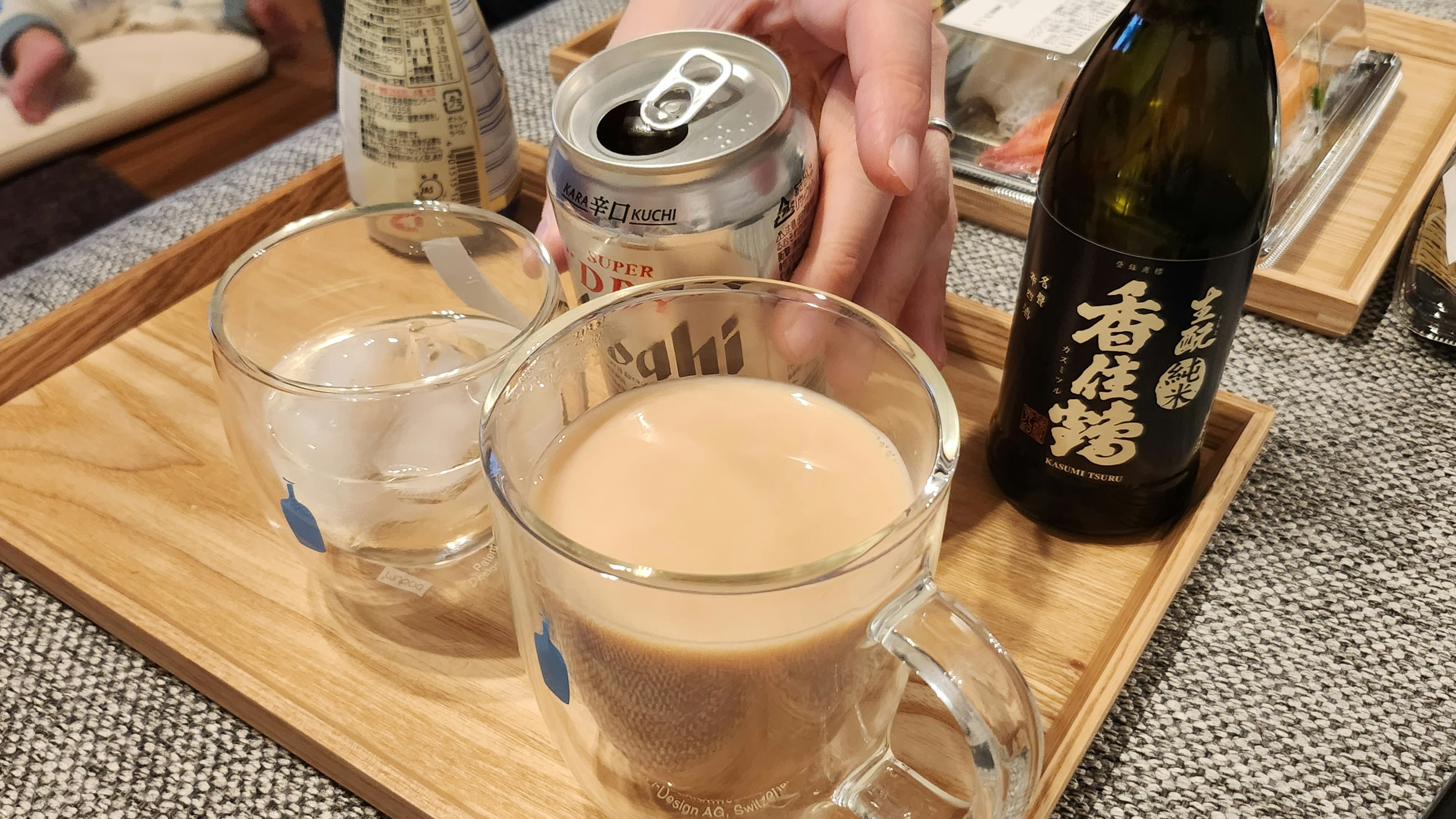 Set minuman di atas nampan kayu dengan gelas transparan berisi es cangkir berisi minuman cokelat dan botol sake hitam