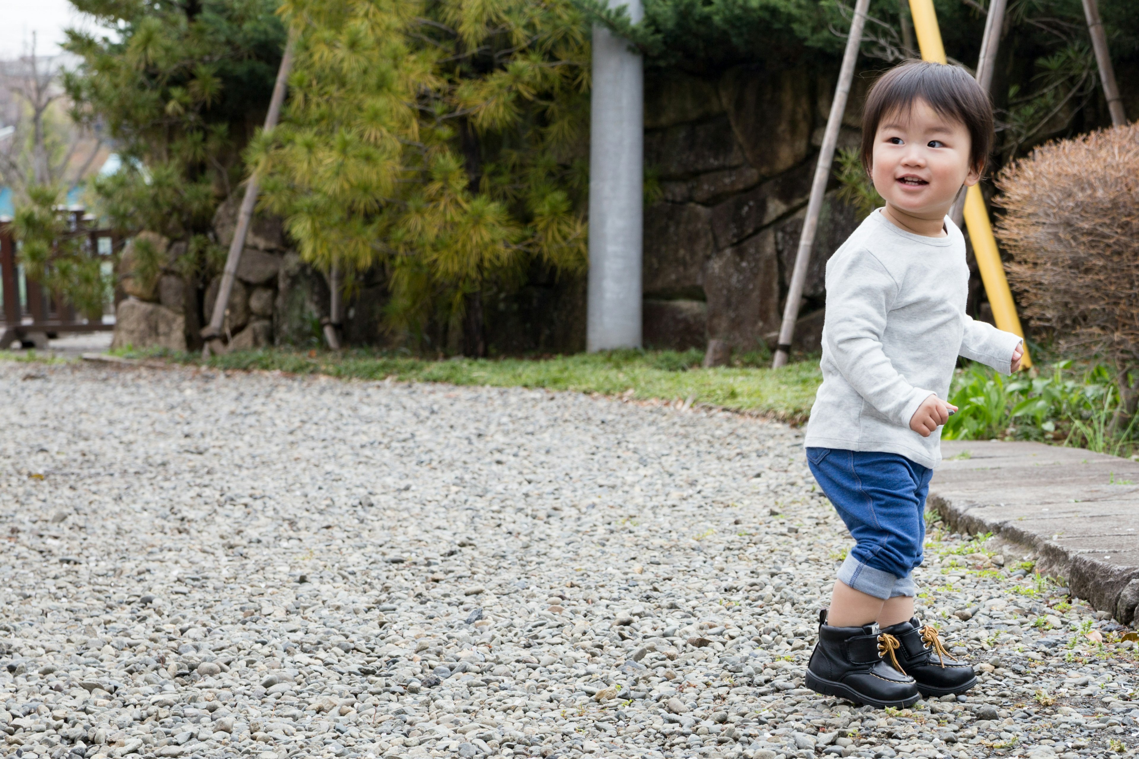 Smiling child playing on a gravel path wearing a white sweater and blue shorts surrounded by green trees