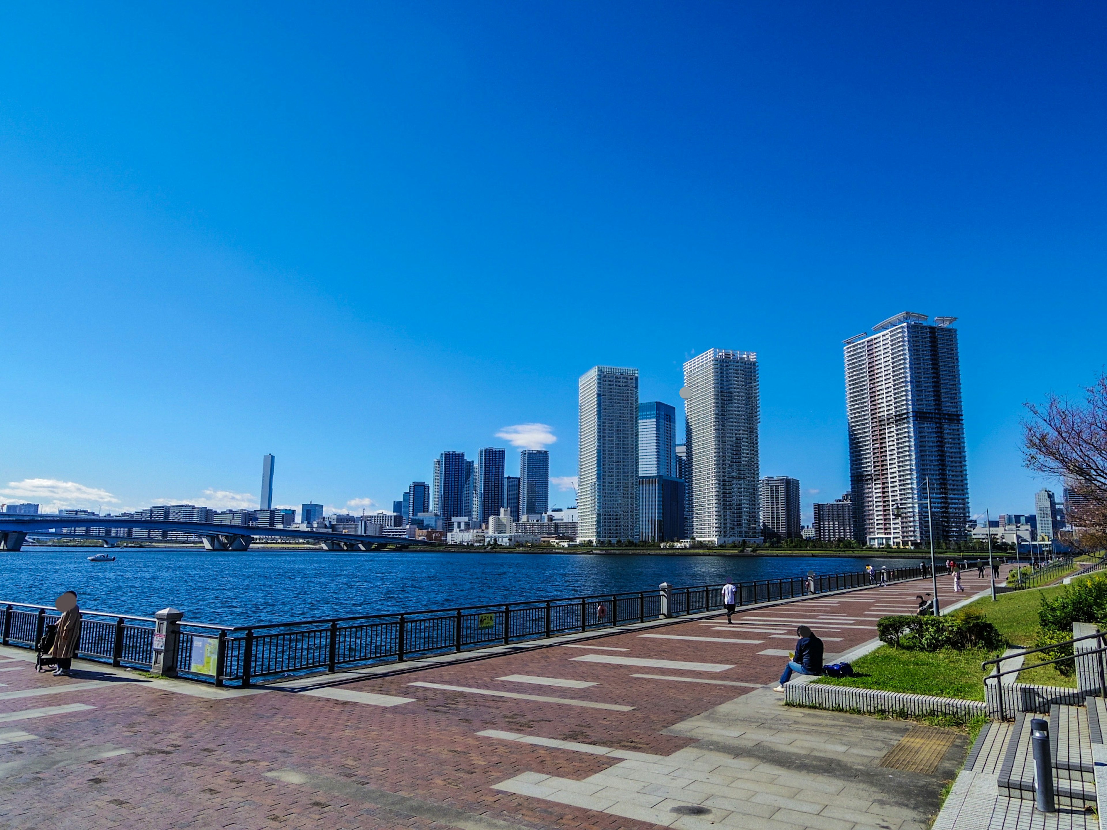 Urban landscape under a blue sky featuring skyscrapers and a river with people on the nearby walkway