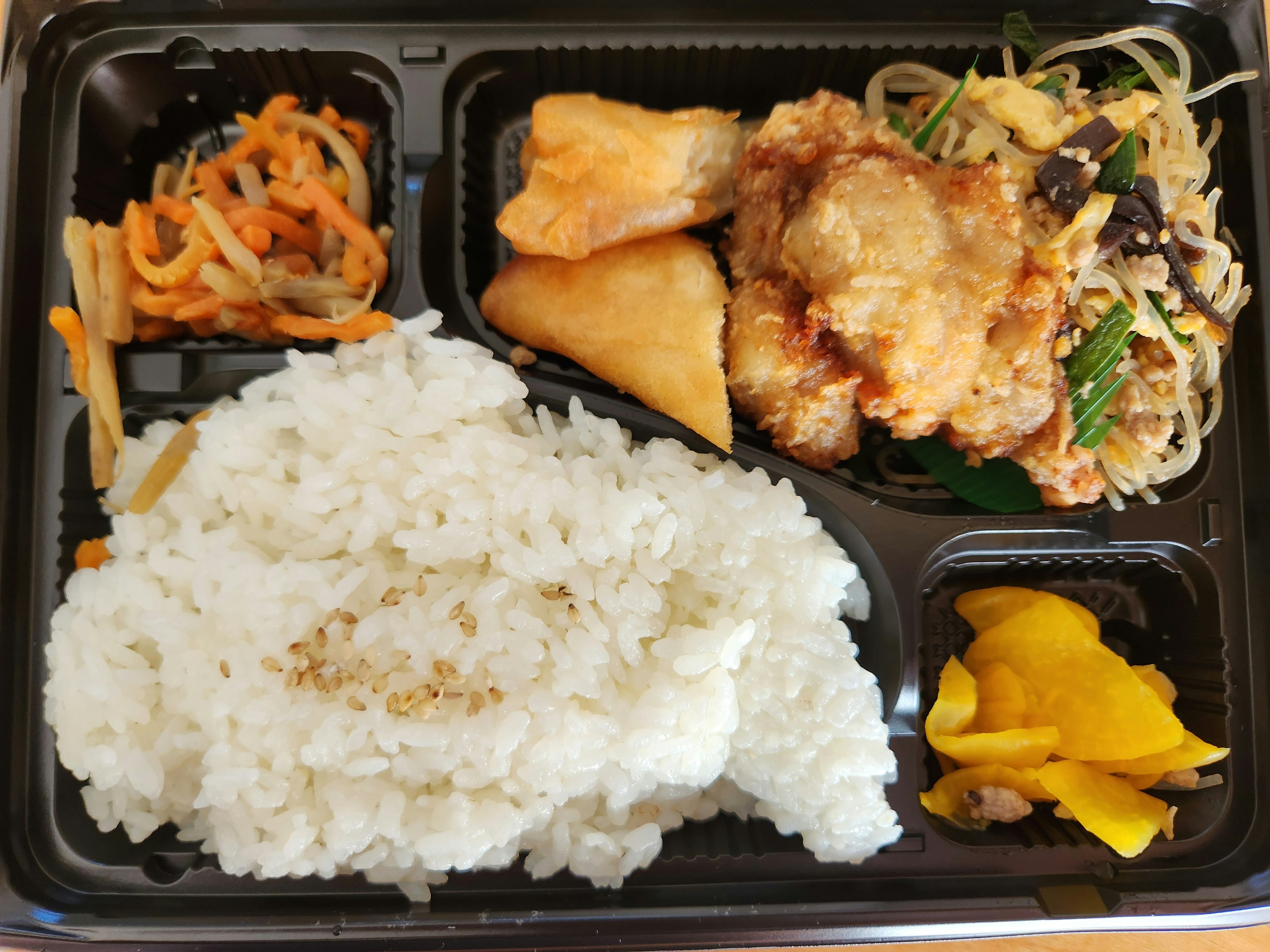 A bento box featuring white rice fried chicken vermicelli salad fried dumplings and vegetable side dishes