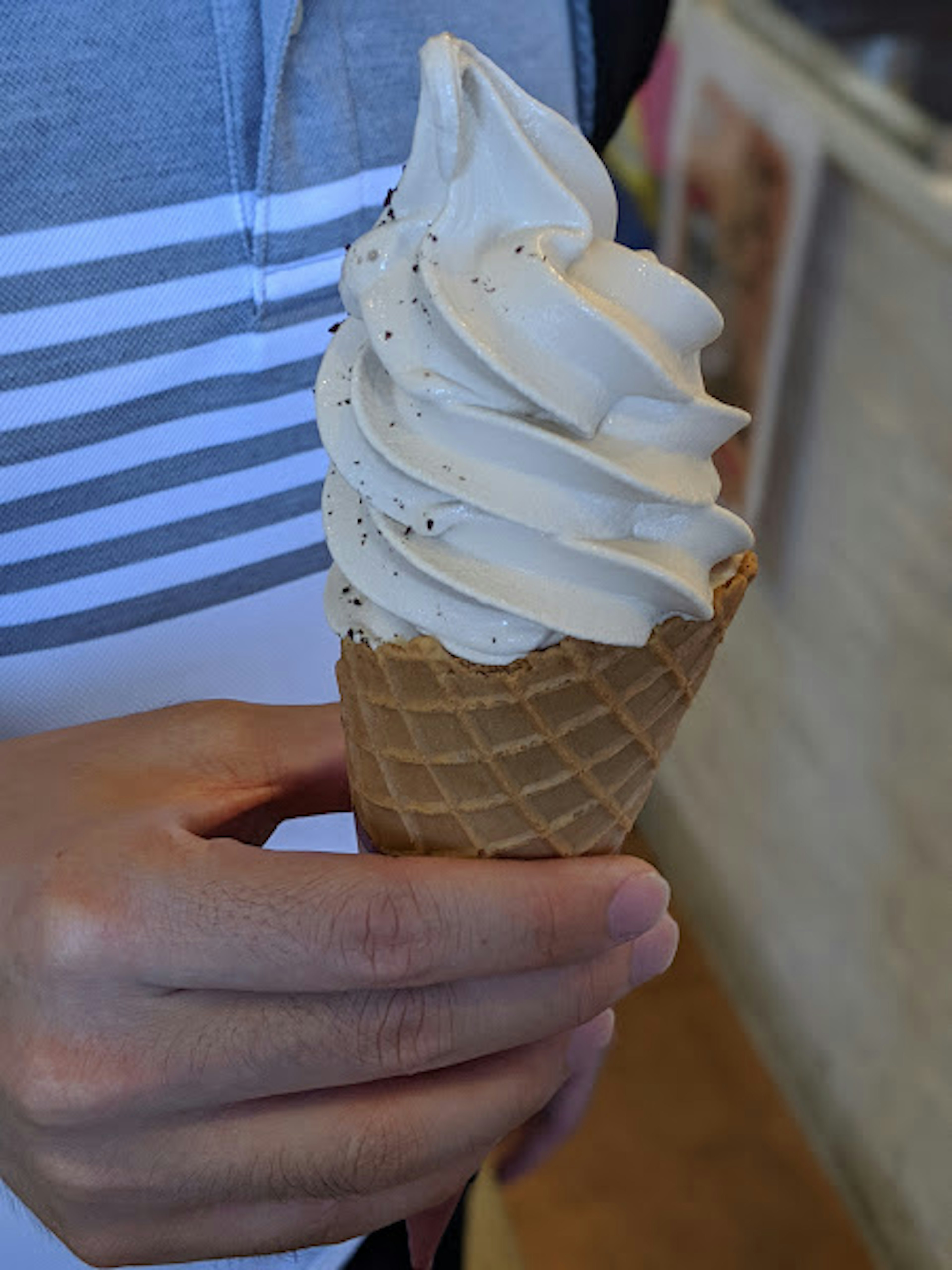 A hand holding a soft serve ice cream in a waffle cone with a creamy color