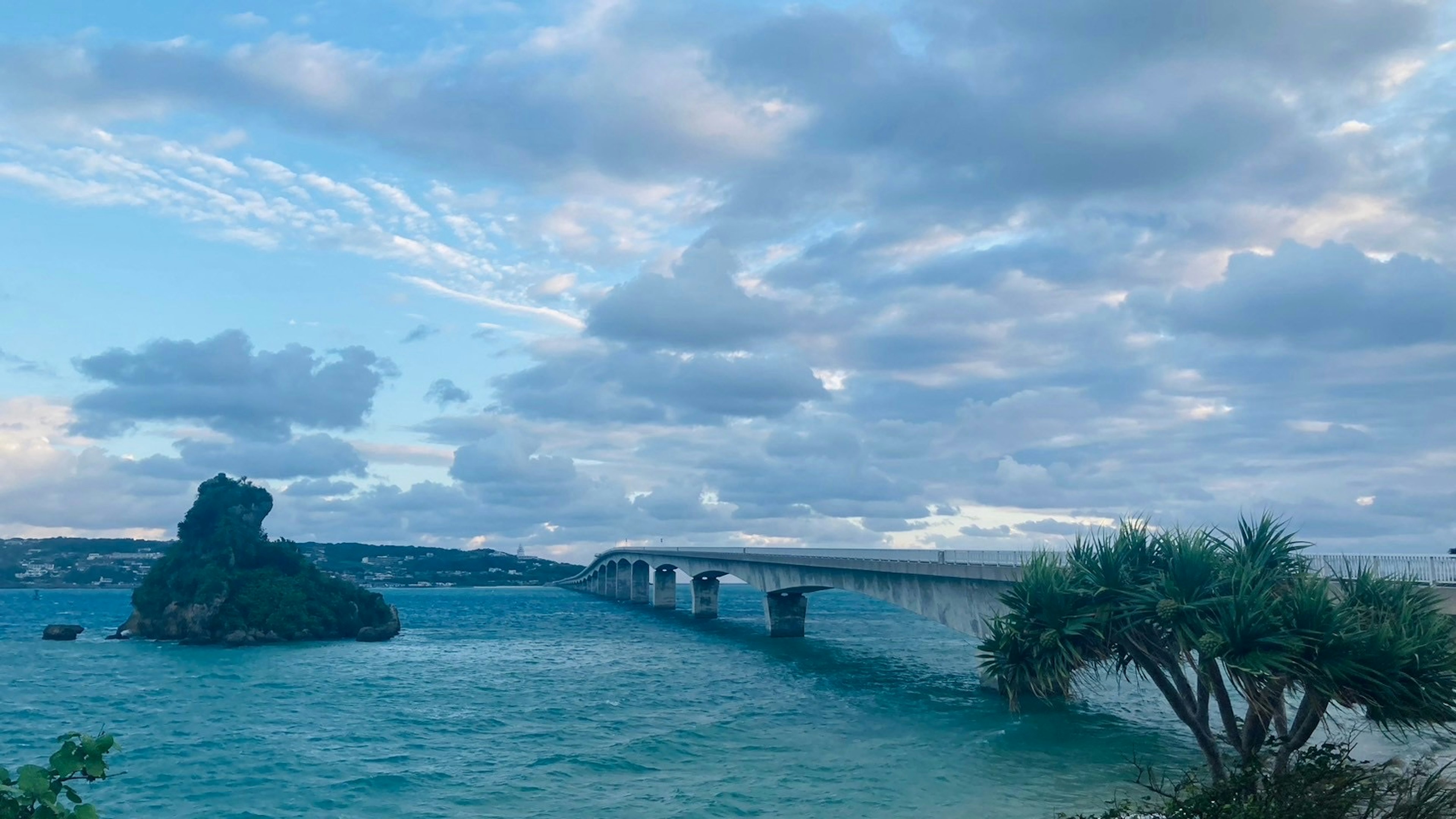 A scenic view of a bridge over blue water with a rock and clouds in the sky