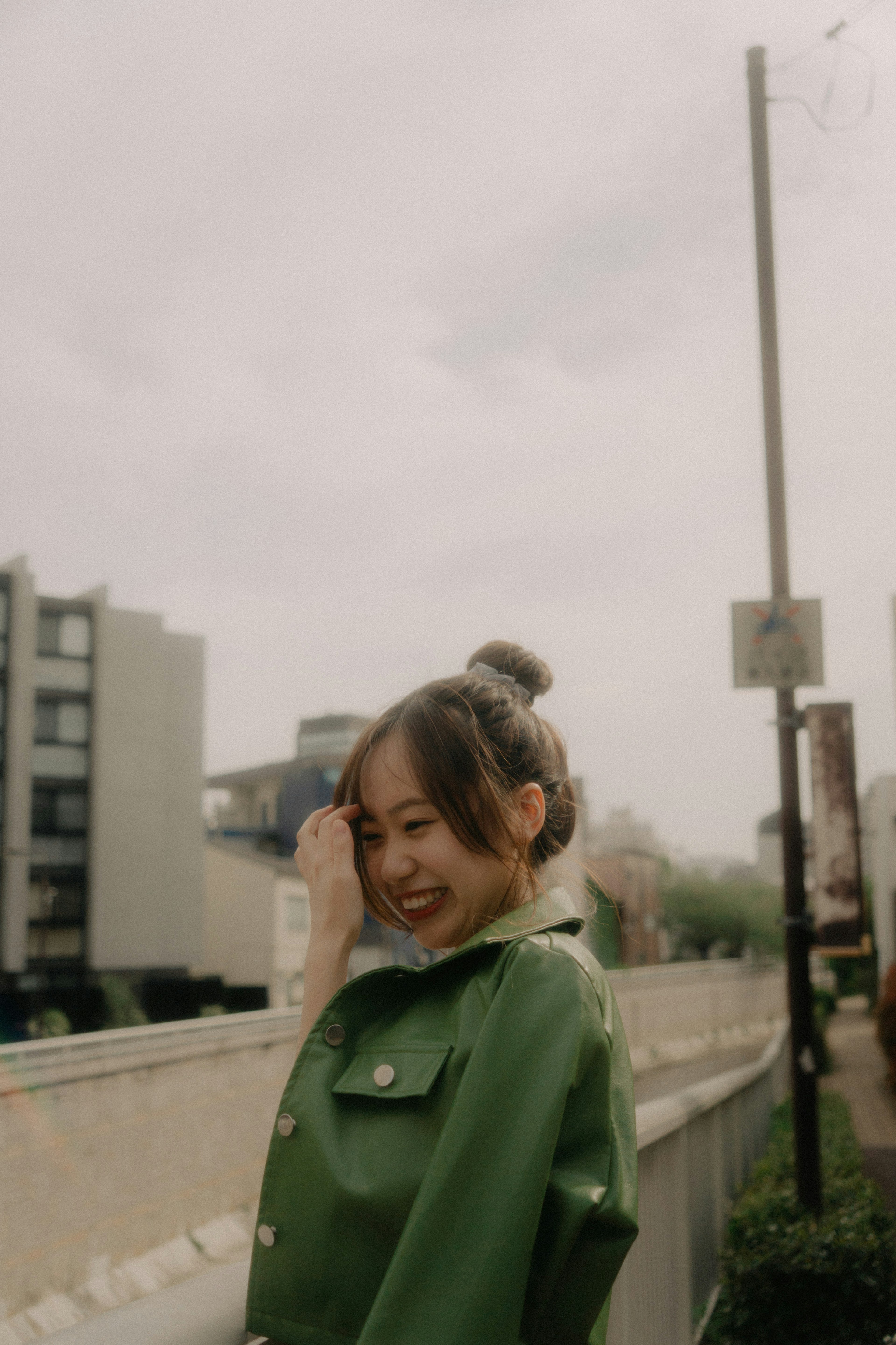 Mujer sonriente con chaqueta verde y fondo urbano bajo un cielo nublado