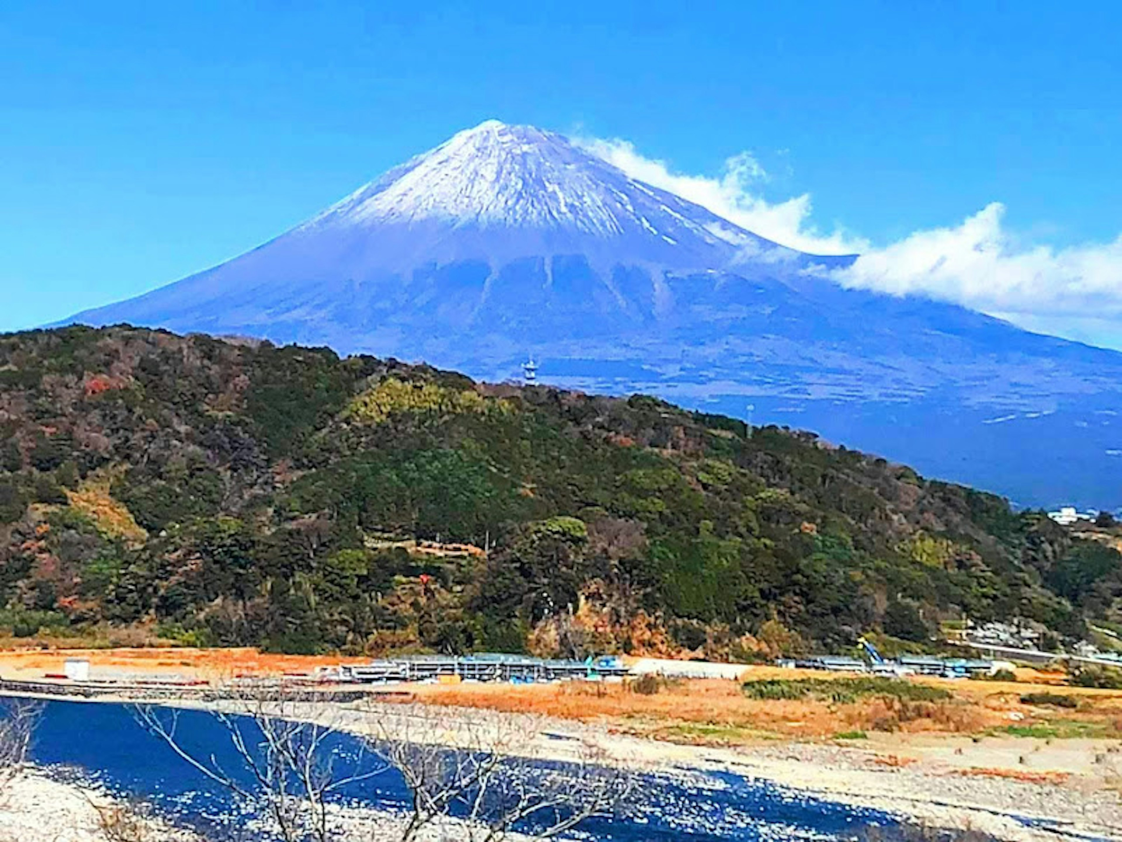 Scenic view of Mount Fuji with a snow-capped peak under a blue sky surrounded by green hills and a river