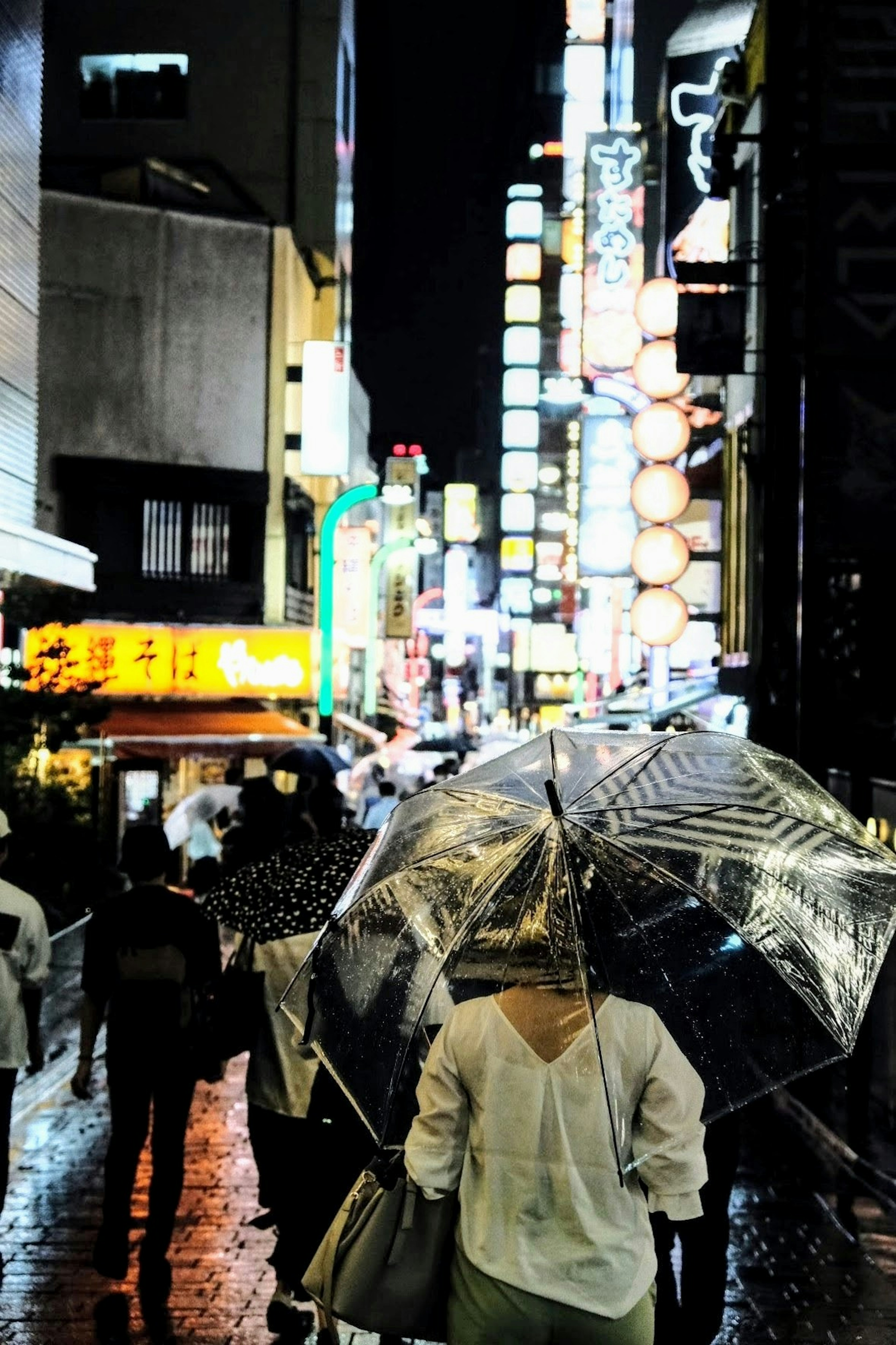 Menschen gehen mit Regenschirmen in einer Stadtstraße bei Nacht Neonlichter reflektieren auf dem nassen Pflaster