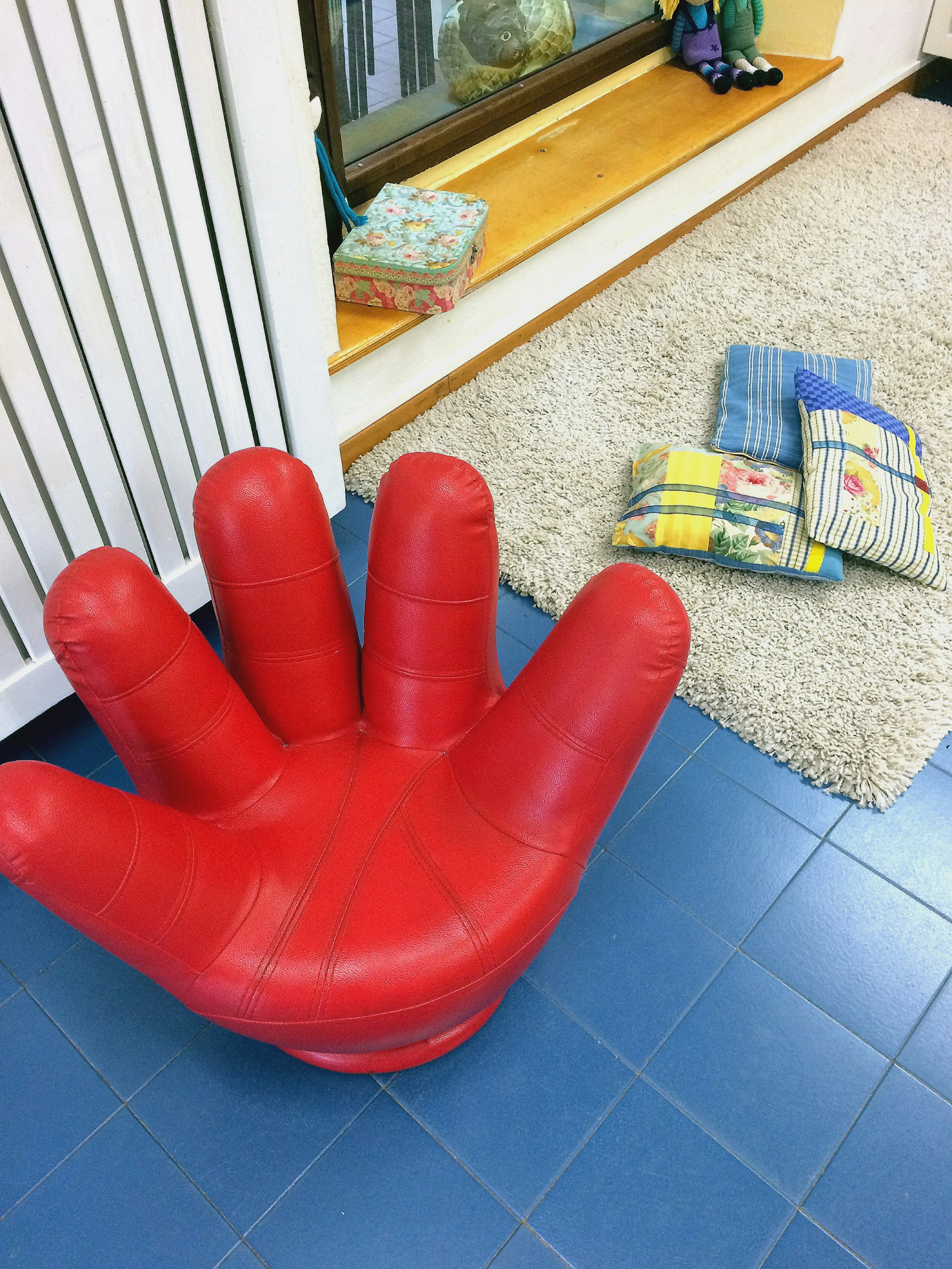 A red hand-shaped chair placed on blue tiles with colorful cushions nearby