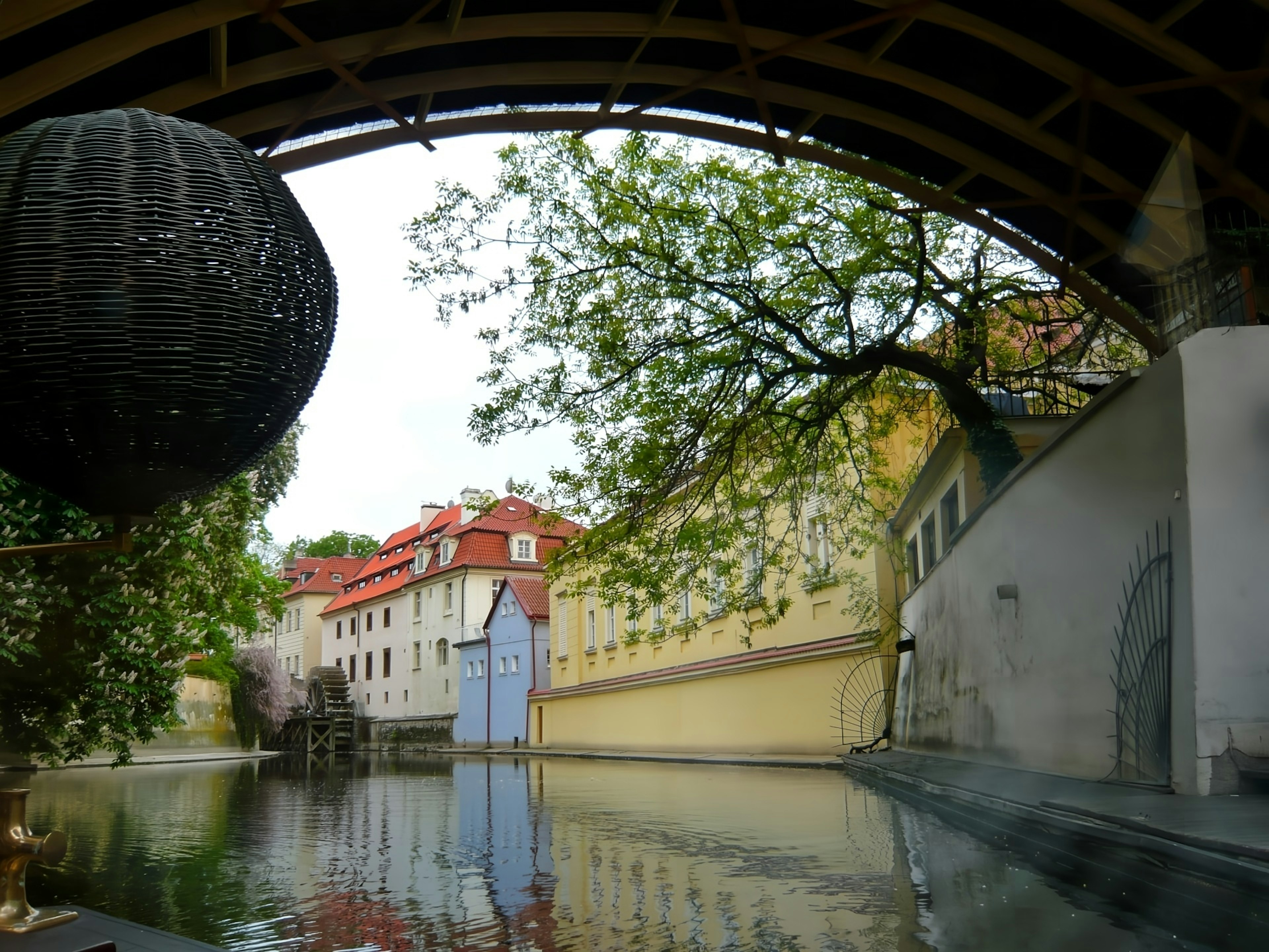 Malersicher Blick von unter einem Bogen auf einen ruhigen Wasserlauf mit Reflexionen von roten Dächern und grünen Bäumen