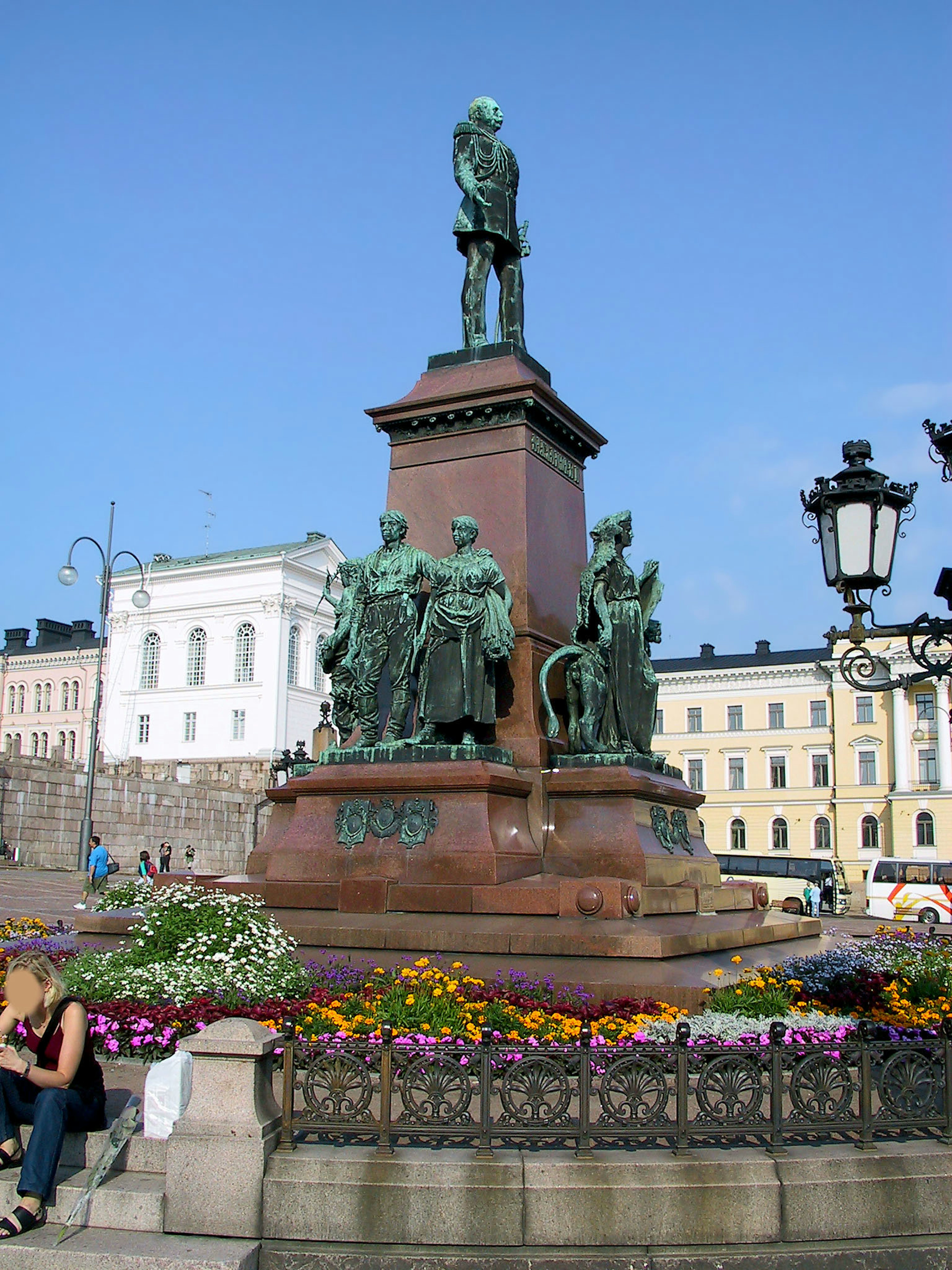 Estatua de bronce en una plaza bajo un cielo azul rodeada de flores