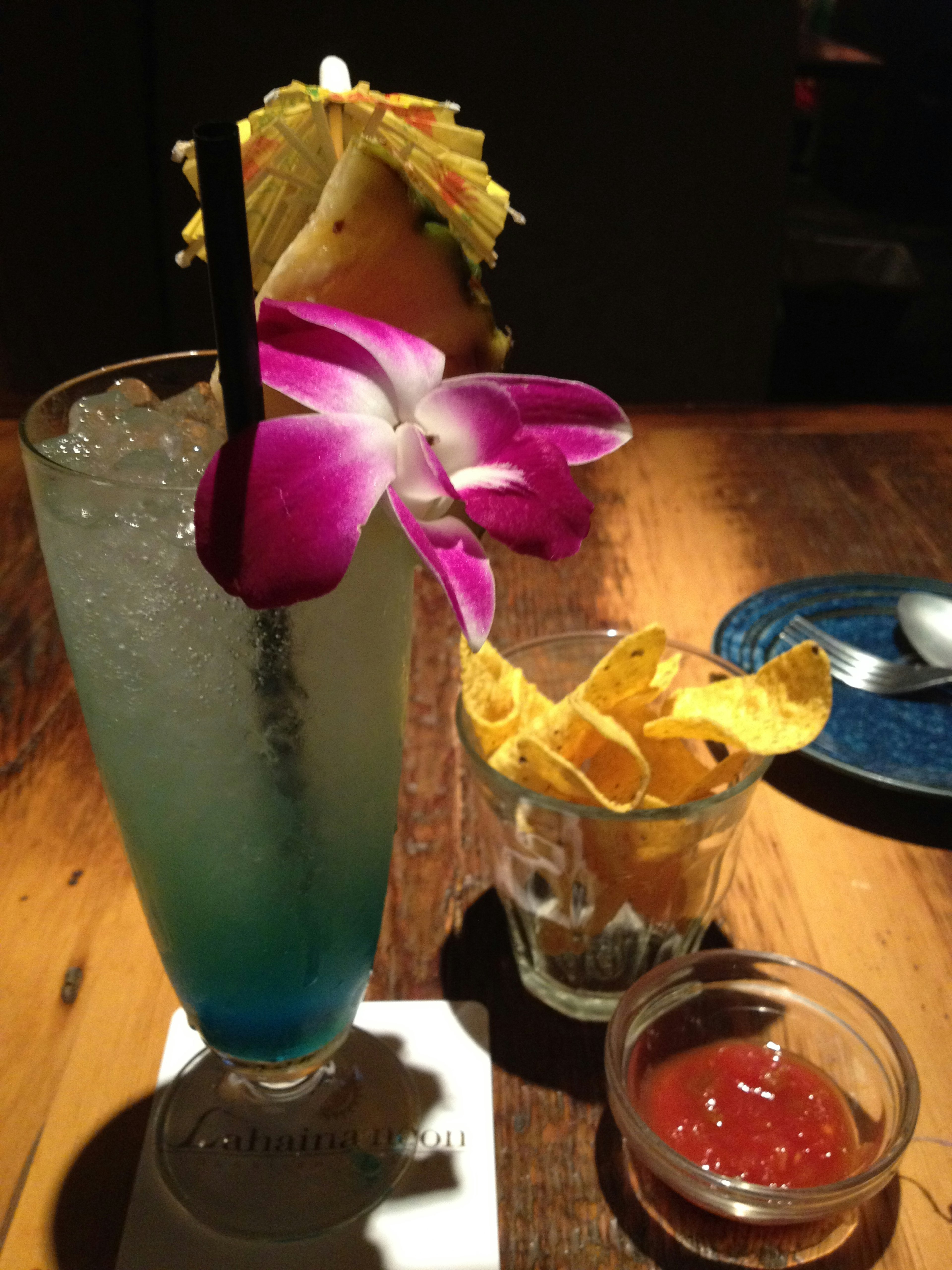 Blue cocktail garnished with a purple flower served with tortilla chips and salsa on a wooden table