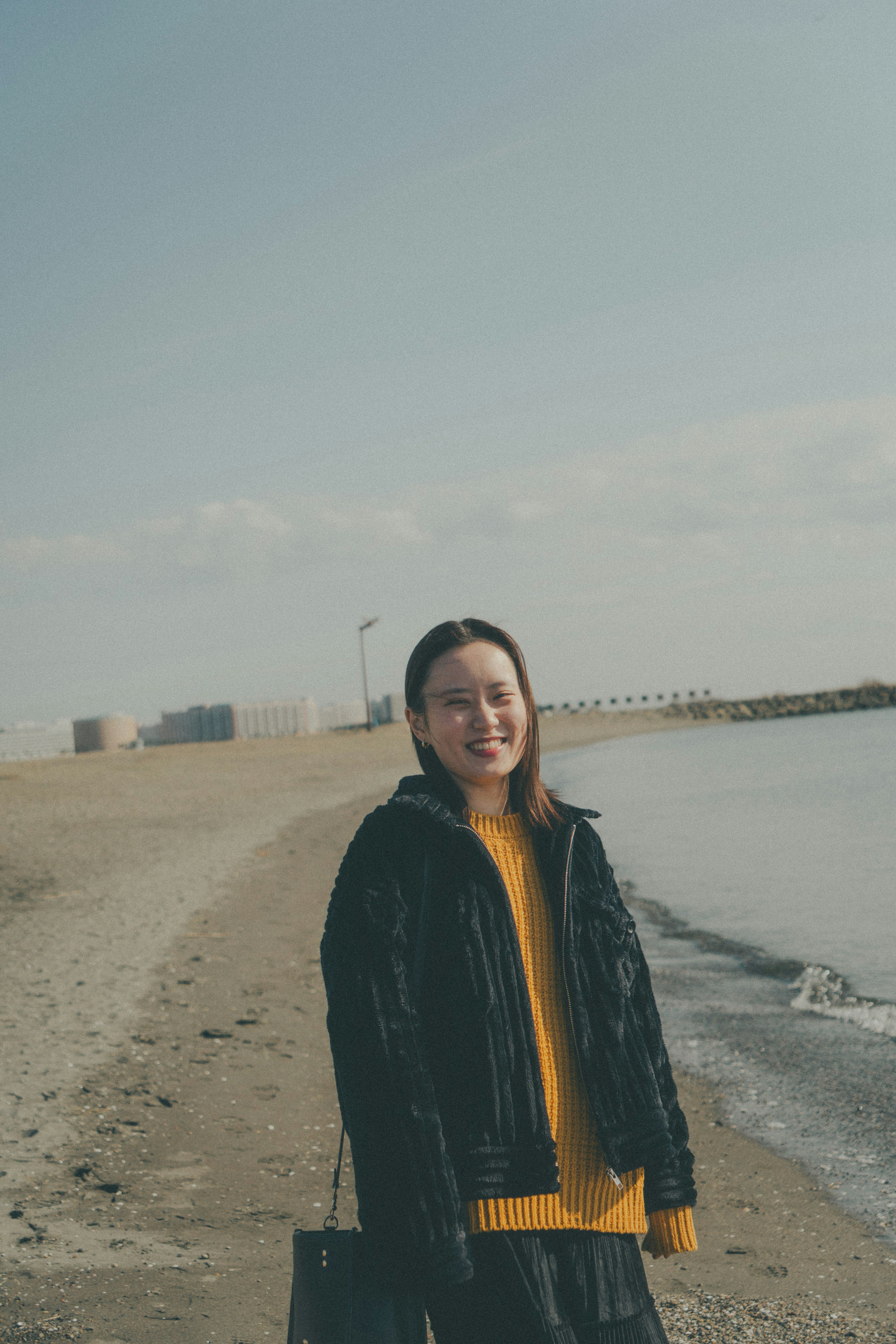 Eine lächelnde Frau am Strand in einer schwarzen Jacke und gelbem Pullover mit Wellen und Sandstrand im Hintergrund